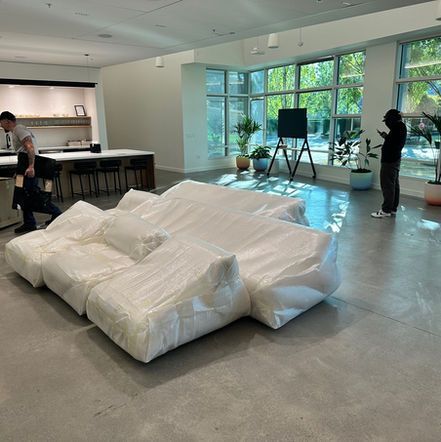 A modern office space features a white sofa covered in plastic, two people, and large windows with plants.