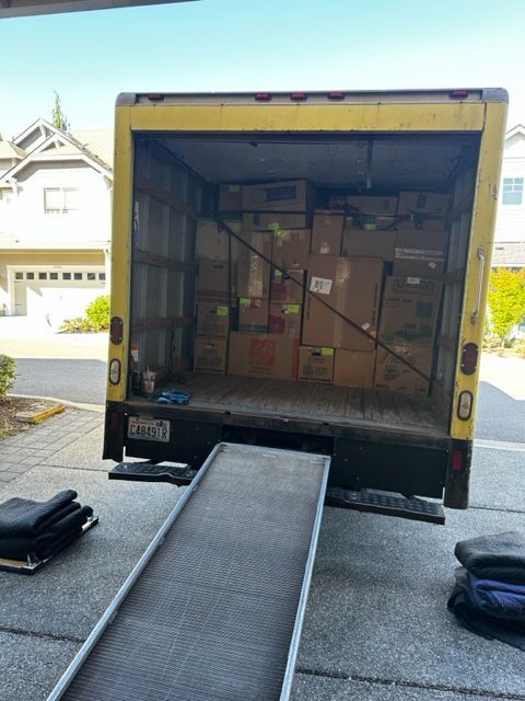Yellow moving truck with ramp extended, full of packed boxes, parked in driveway.