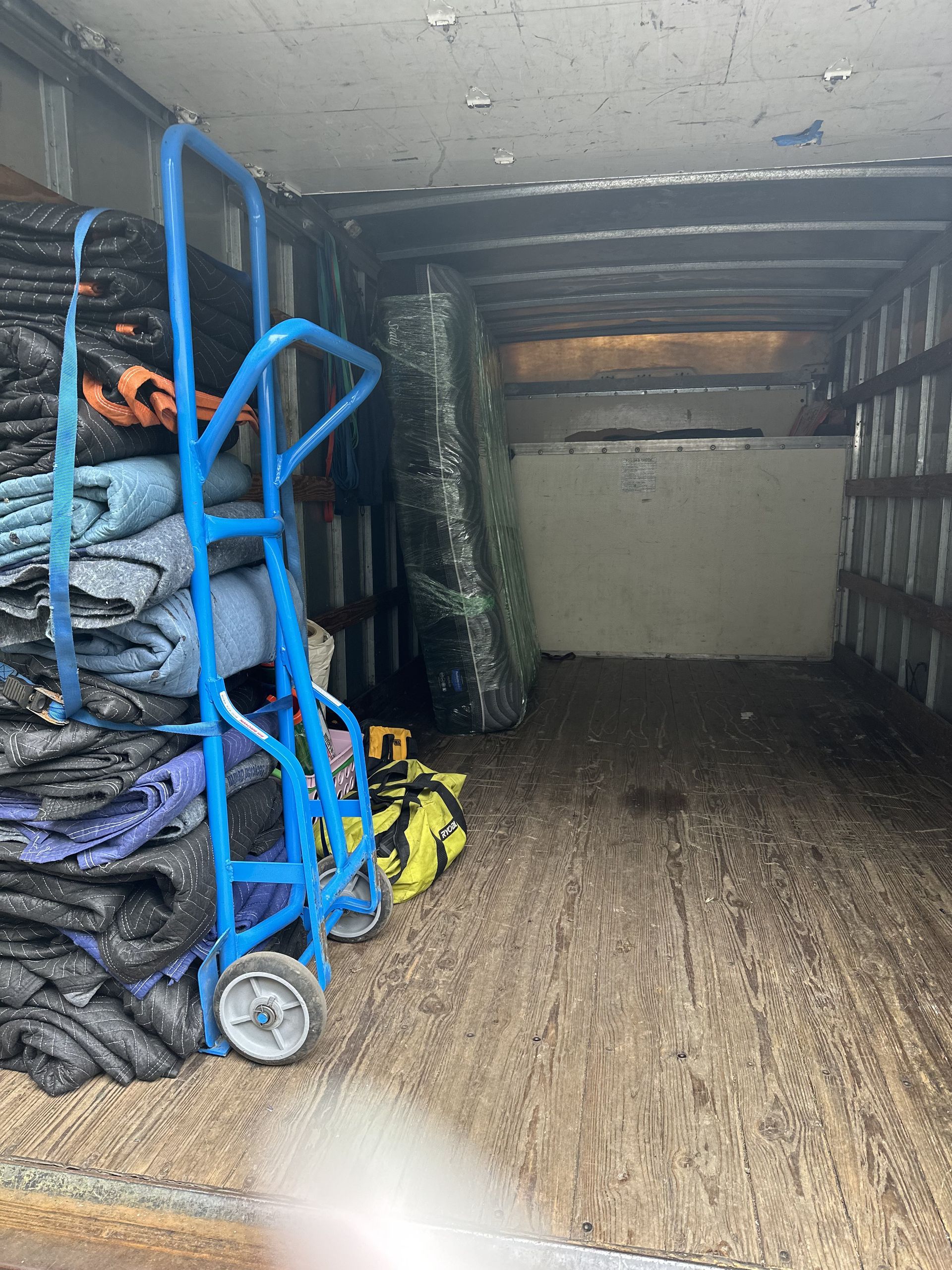 Inside a cargo truck: a blue dolly, stacked furniture pads, wrapped items, and a large mattress.