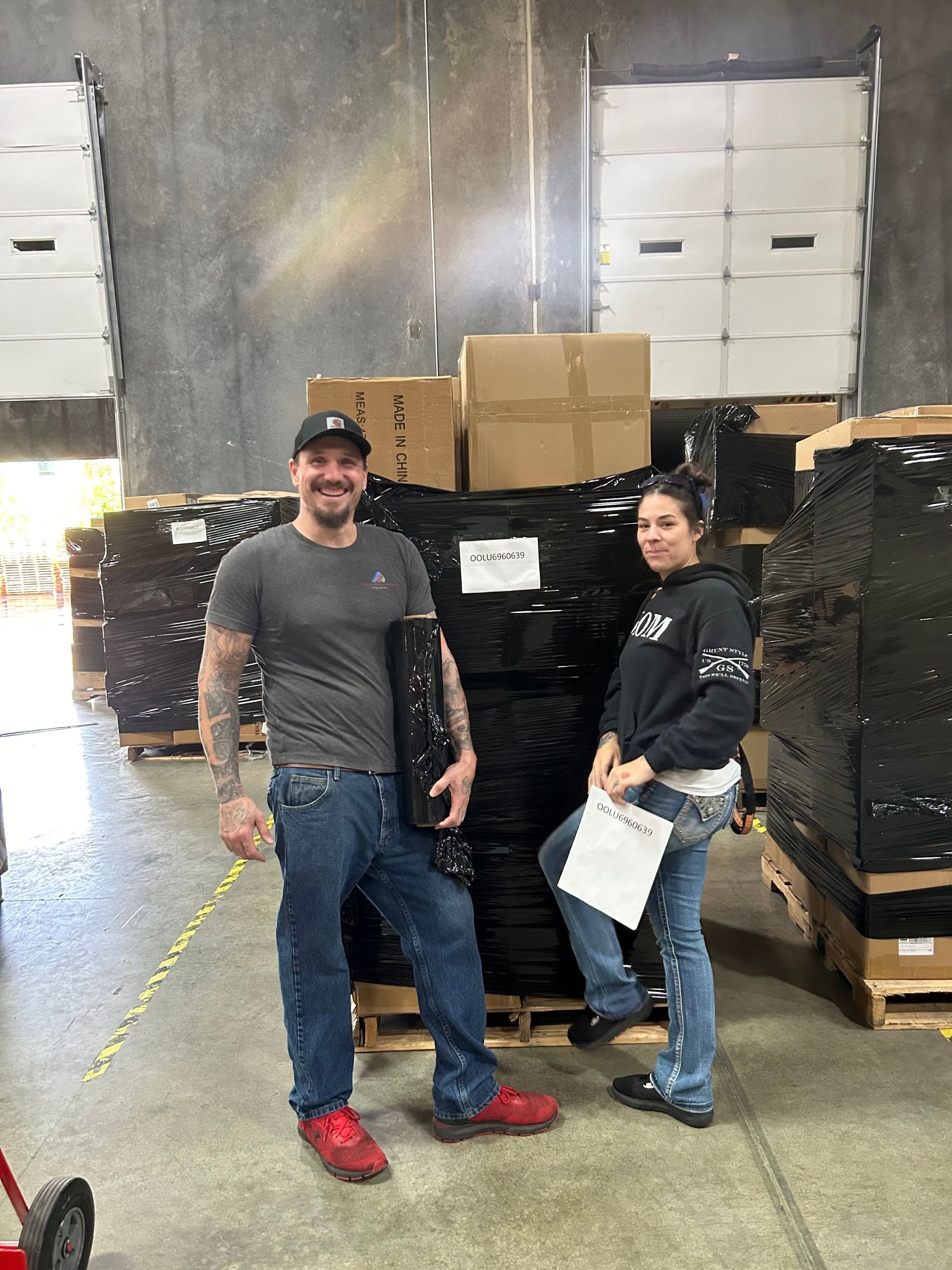 Two people in warehouse stand by pallets of black items, boxes, and a pallet jack.
