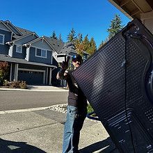 Man carrying large black object outdoors, houses in the background, sunny day.