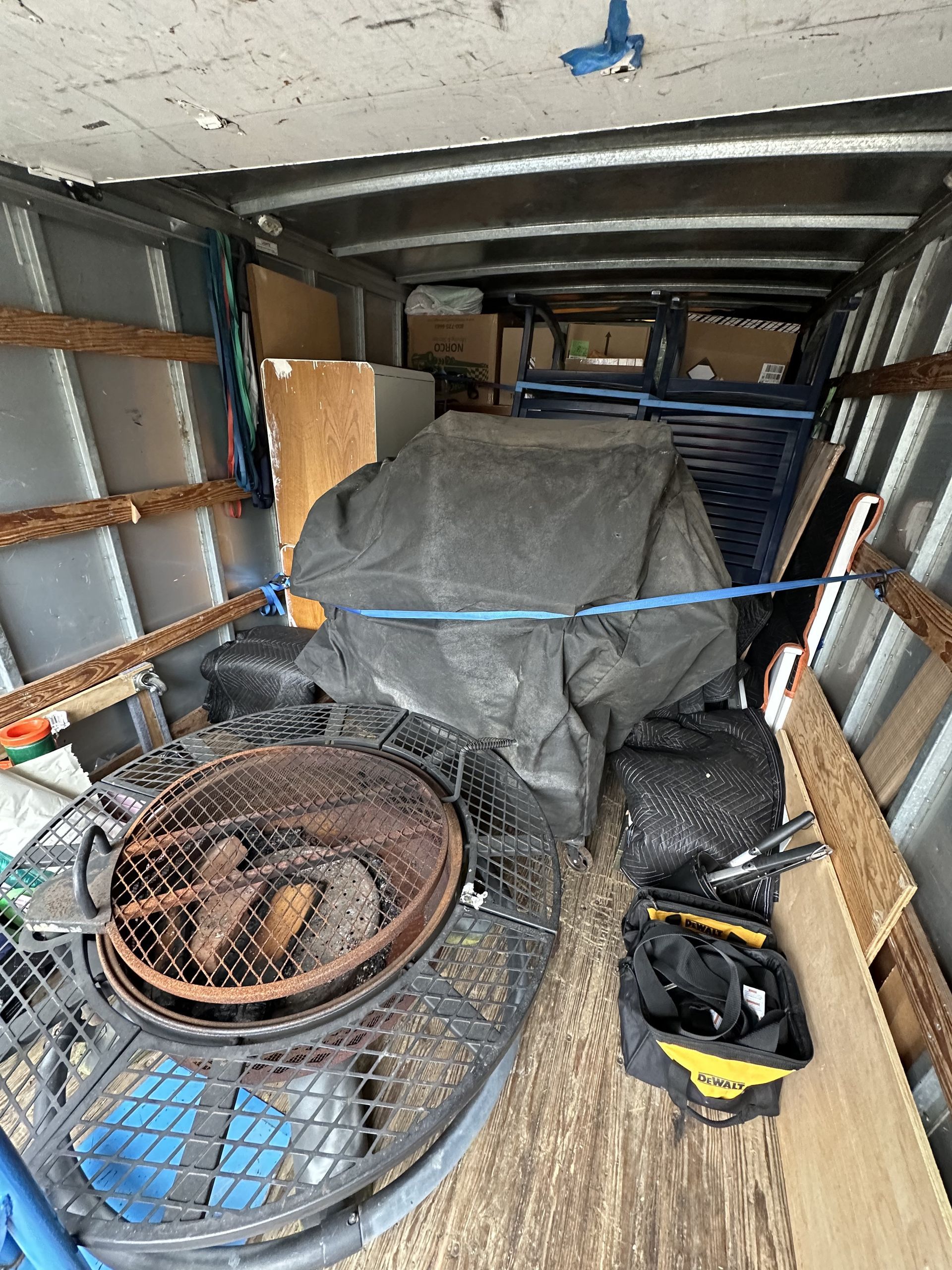 Inside of a truck bed, loaded with various items, including a grill, covered objects, and a tool bag.