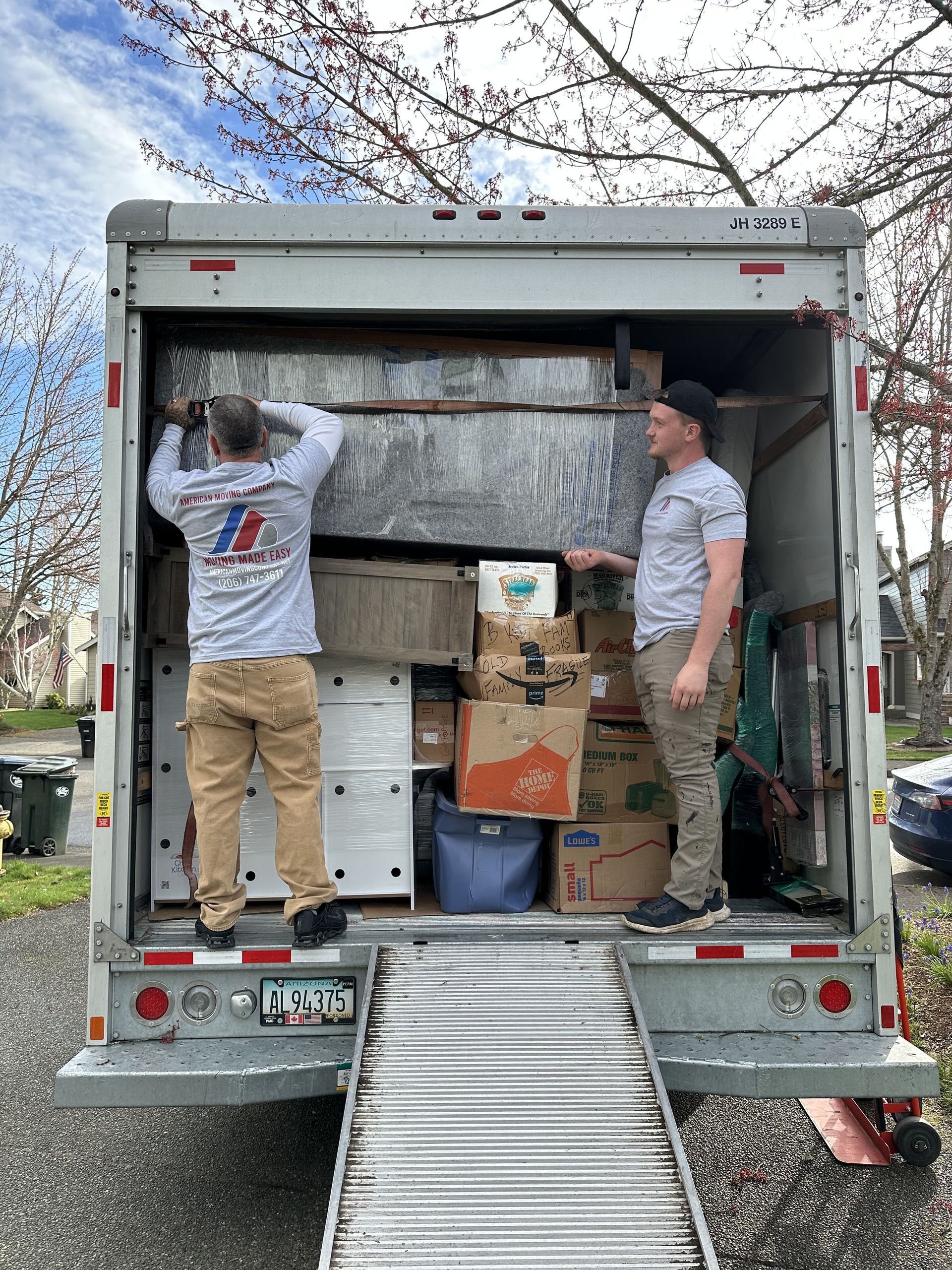 Two movers loading boxes and furniture into a moving truck with ramp outdoors.