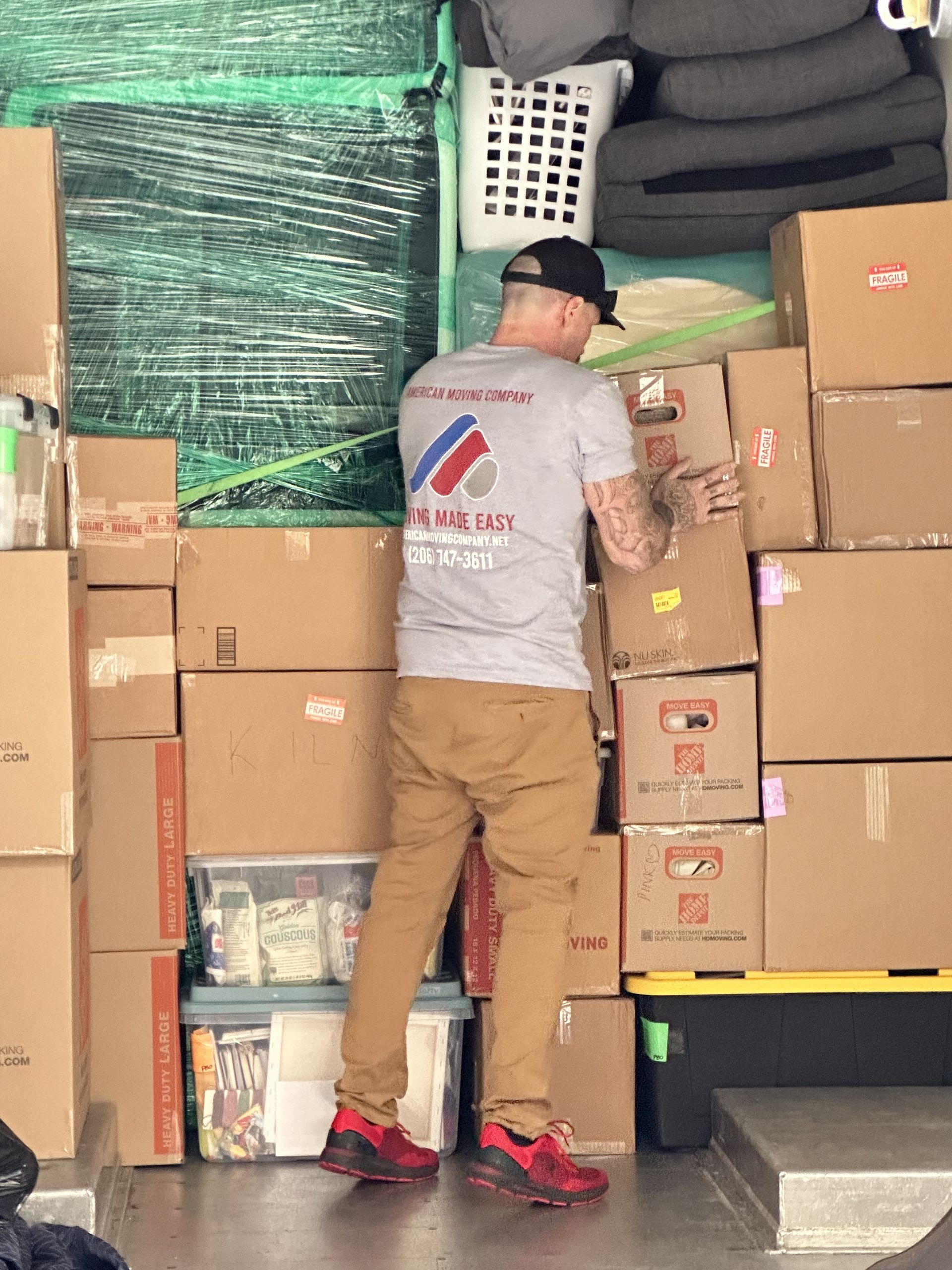 A person stacks boxes inside a moving truck. The person wears a t-shirt, brown pants, and red shoes.