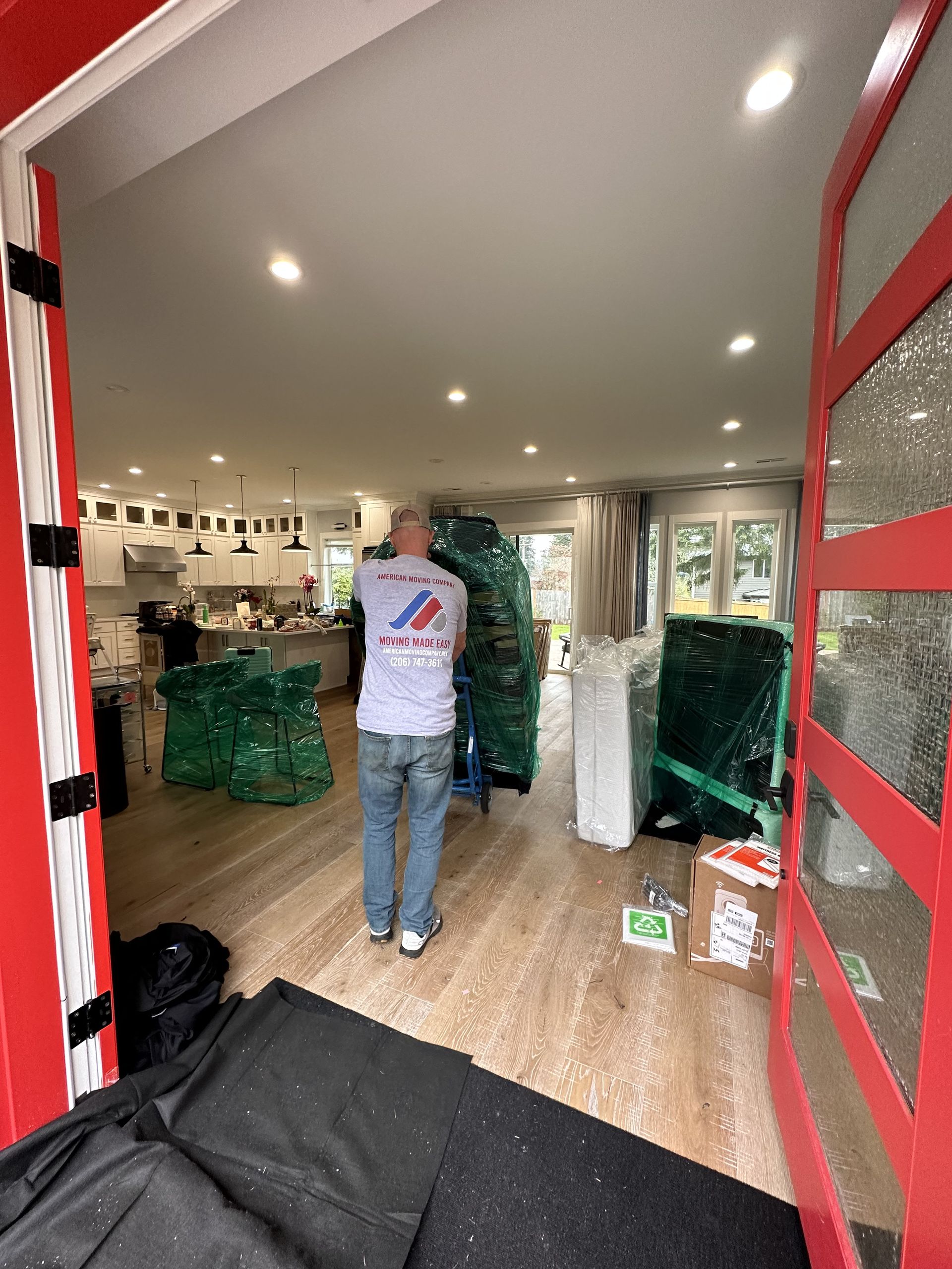 Man pushing a dolly loaded with wrapped furniture into a brightly lit house. Red door frame.