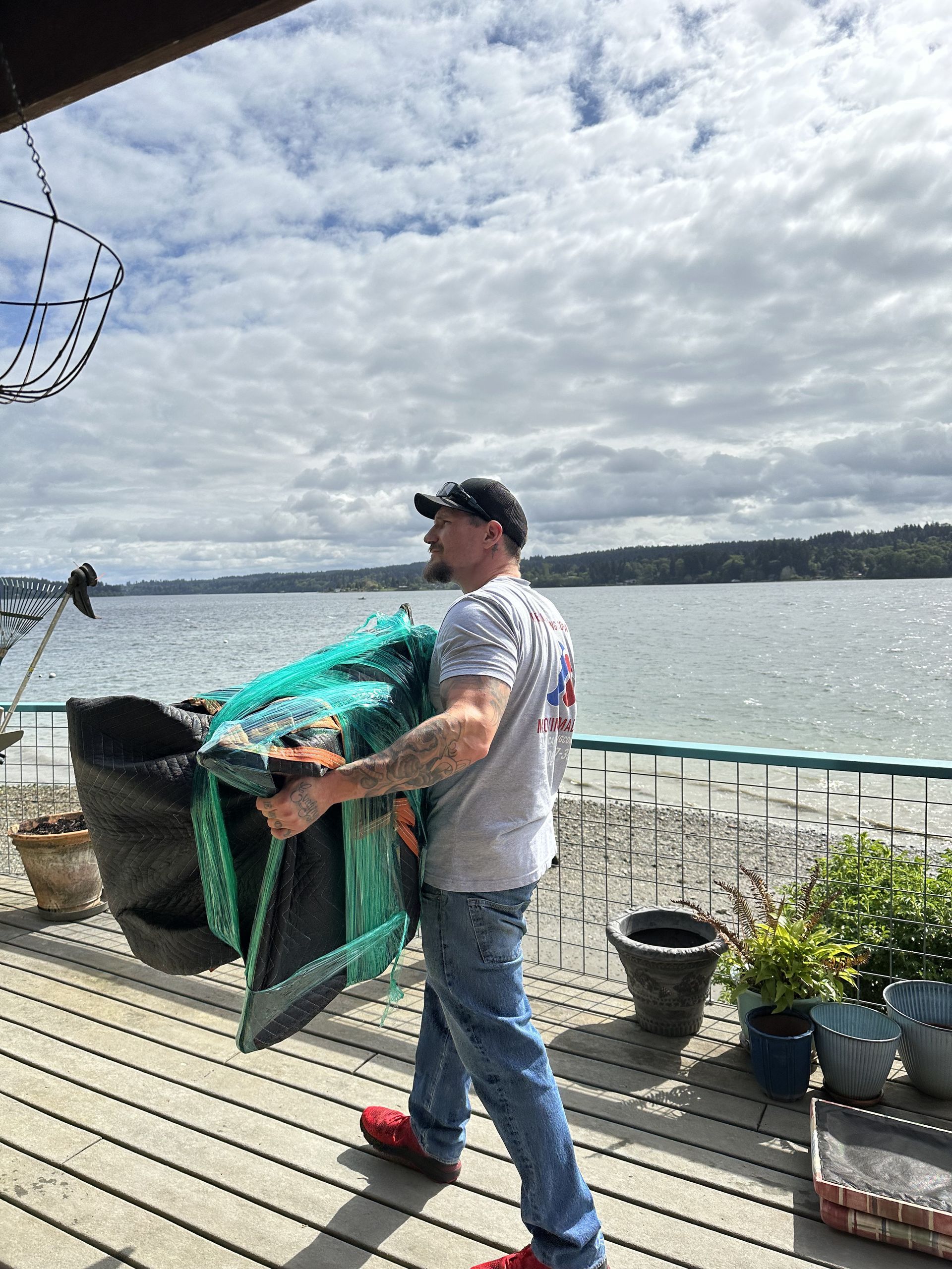 Man carrying a large green and black object on a waterfront deck, cloudy sky and water in the background.