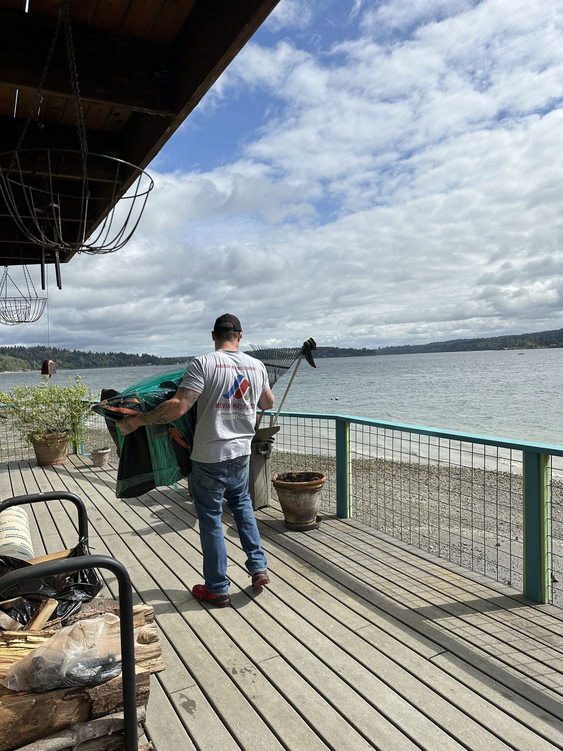 Man on wooden deck carrying blanket towards water. Cloudy sky.