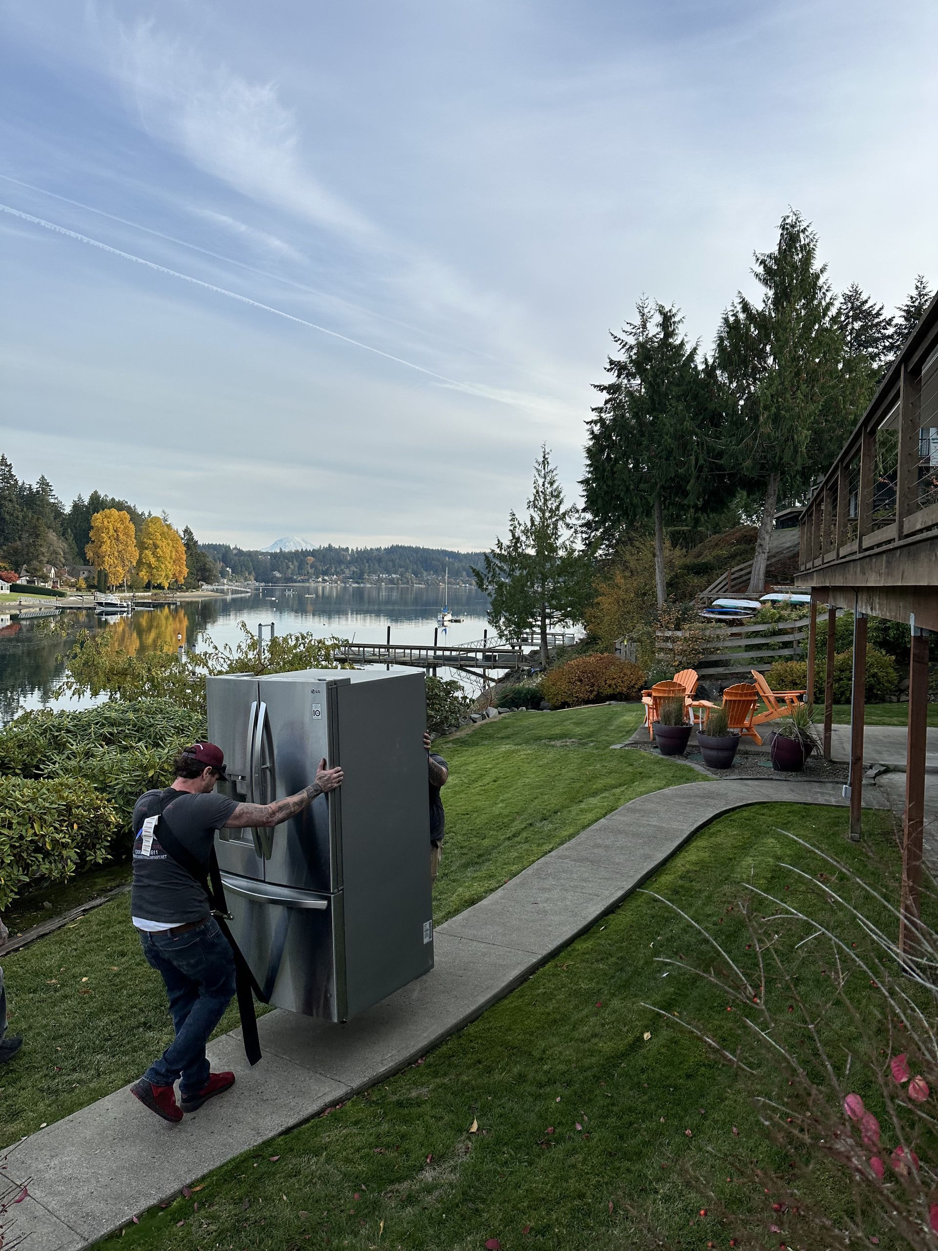 Person carrying a refrigerator on a pathway beside a lake with trees.