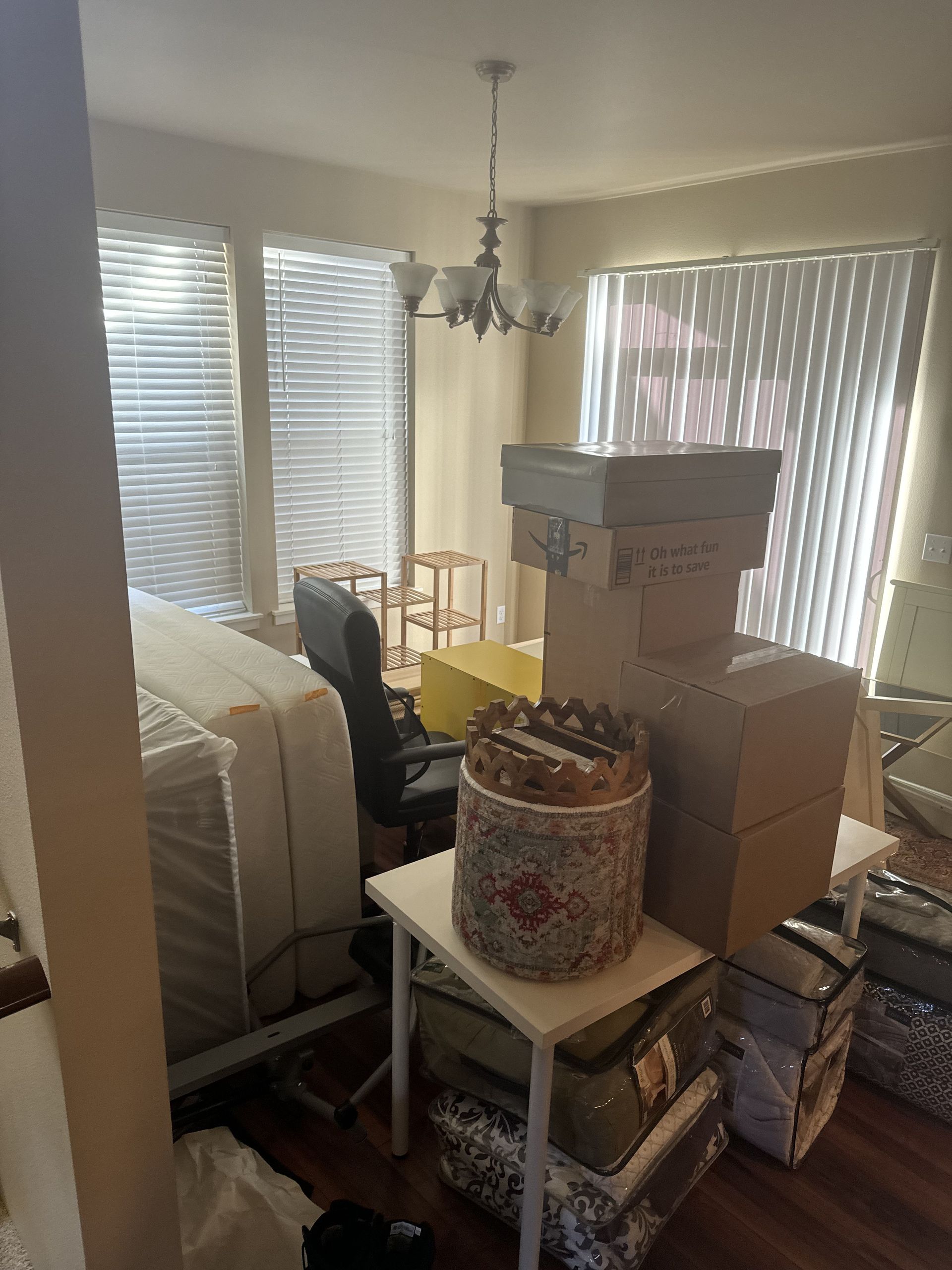Room with boxes, a bed, desk, and chandelier. Blinds cover the windows.