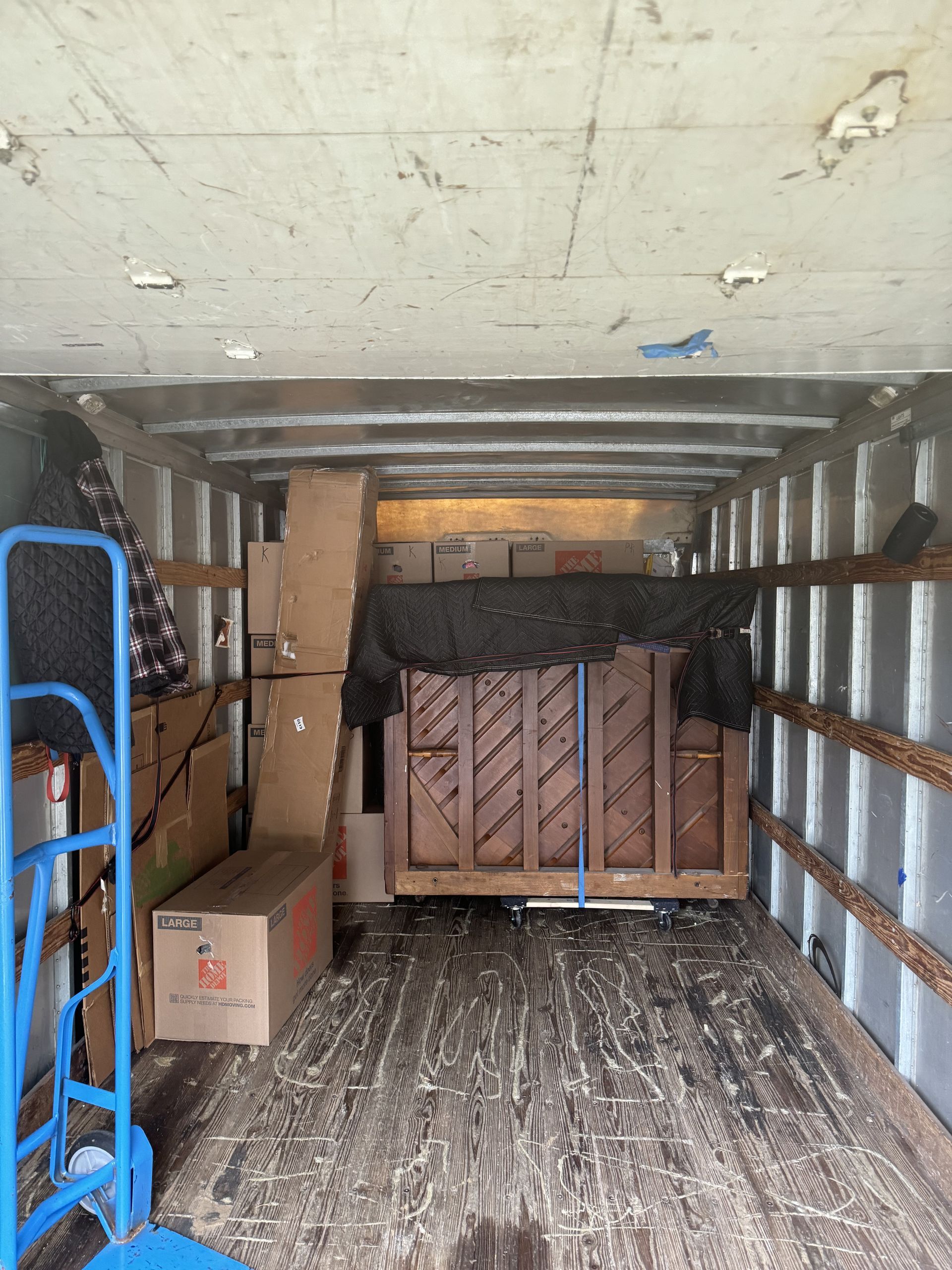 Inside of a moving truck: a piano secured on a wooden platform, surrounded by boxes and other items.