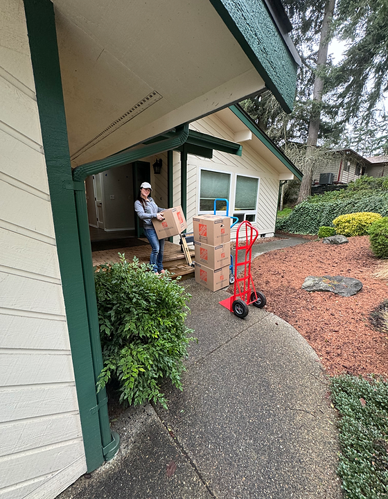 Person moving boxes outside a house; a red dolly with boxes sits nearby.