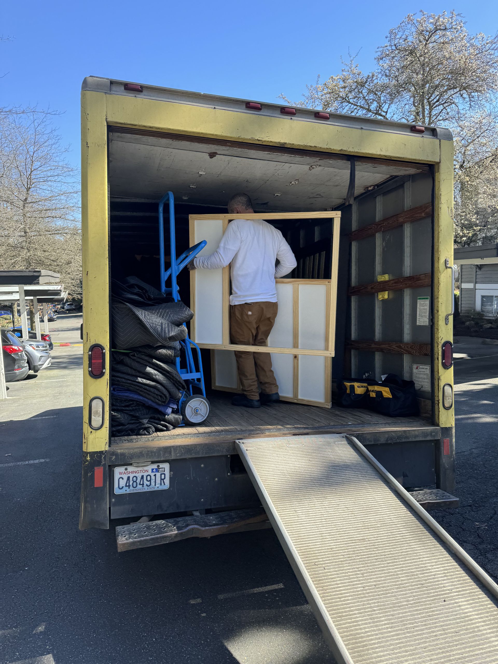 Person loading a large, rectangular framed object into a yellow truck with a ramp extended.
