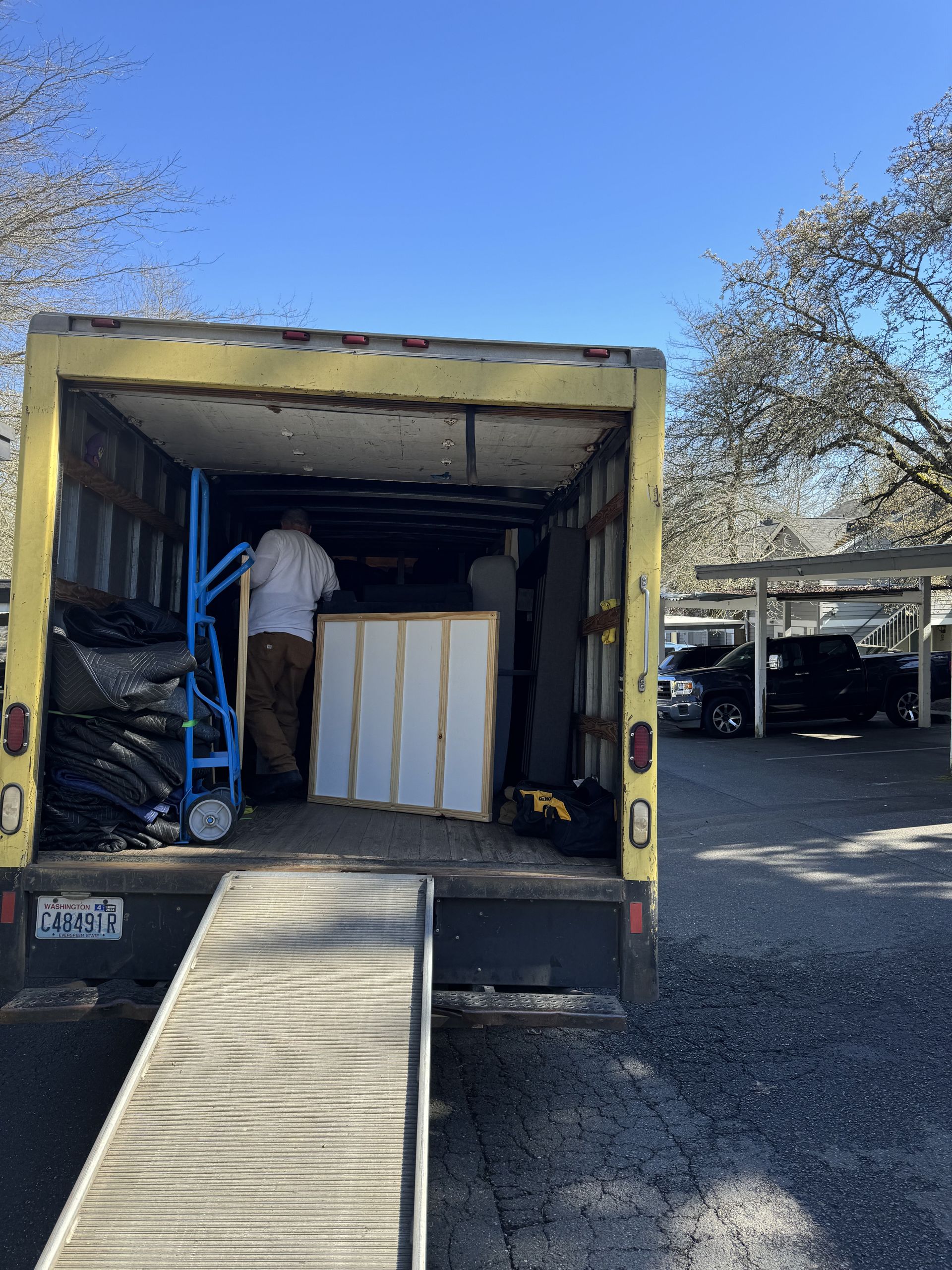 Yellow moving truck open with people loading items onto the ramp, parked outside under a blue sky.