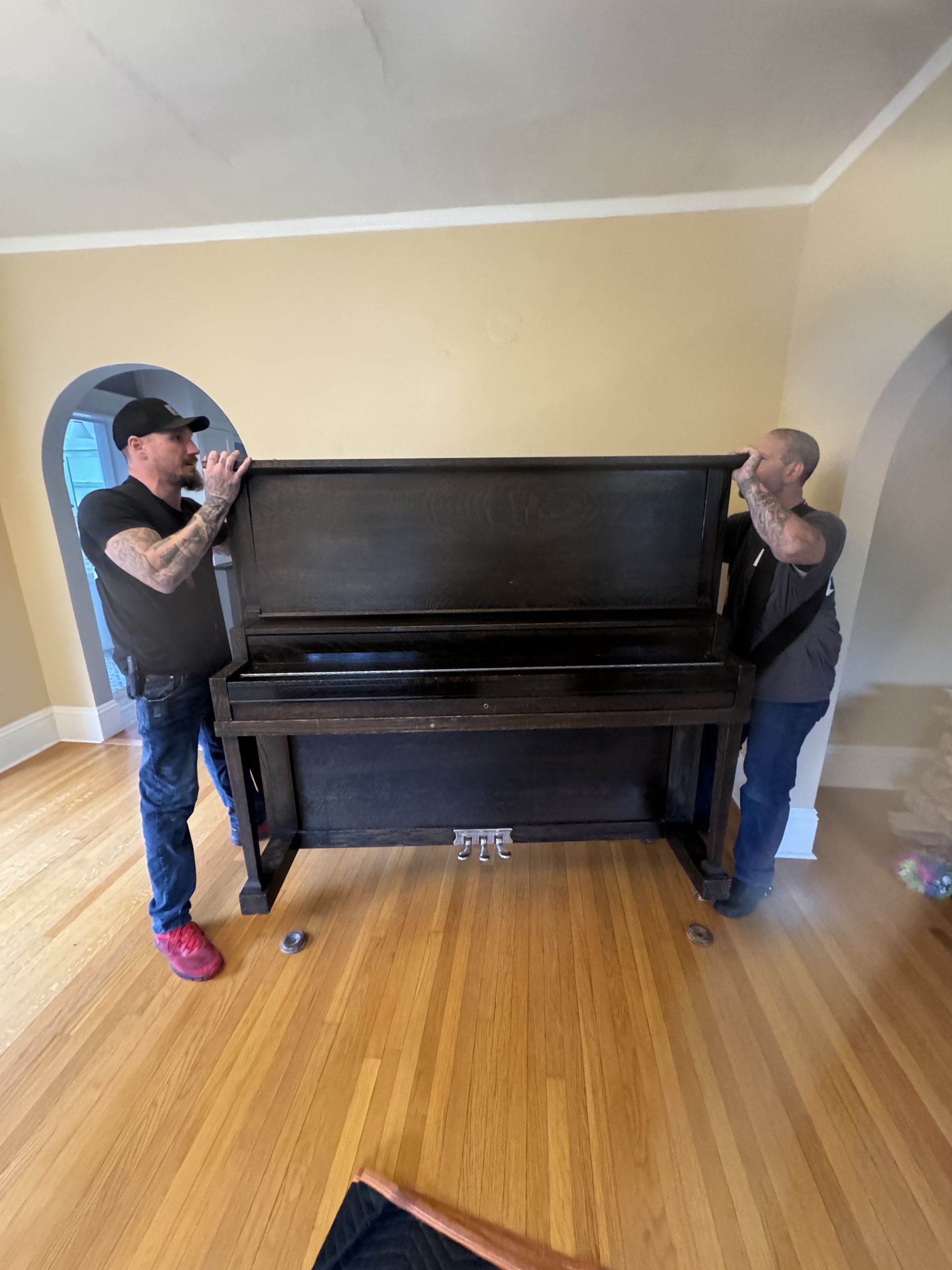 Two people lifting a dark upright piano in a room with hardwood floors and tan walls.