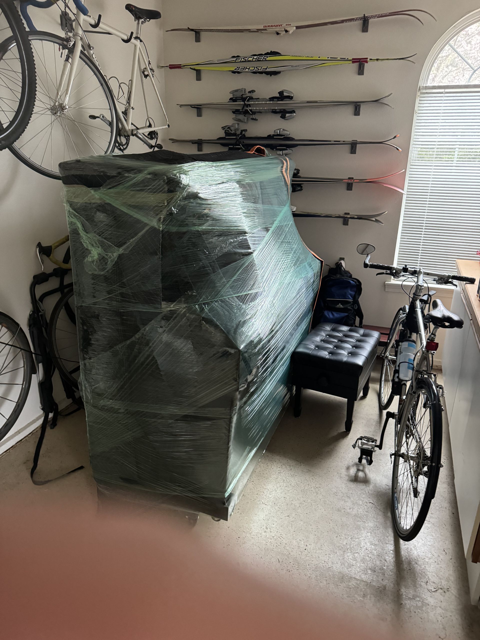 Piano wrapped in plastic, with a piano bench, skis, and bikes in a garage.