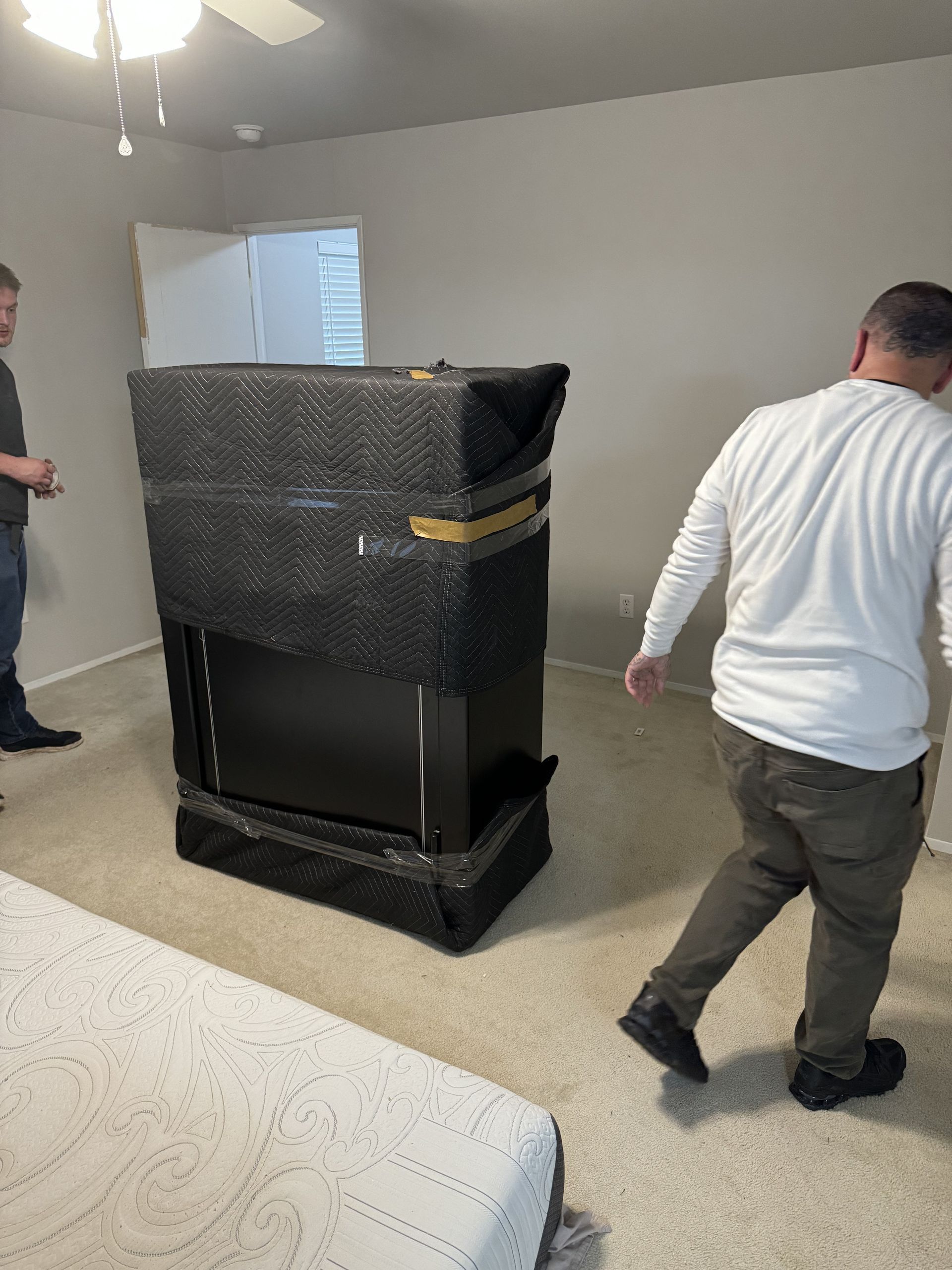 Two people moving a large, black, wrapped object in a beige-carpeted room. A bed is in the foreground.