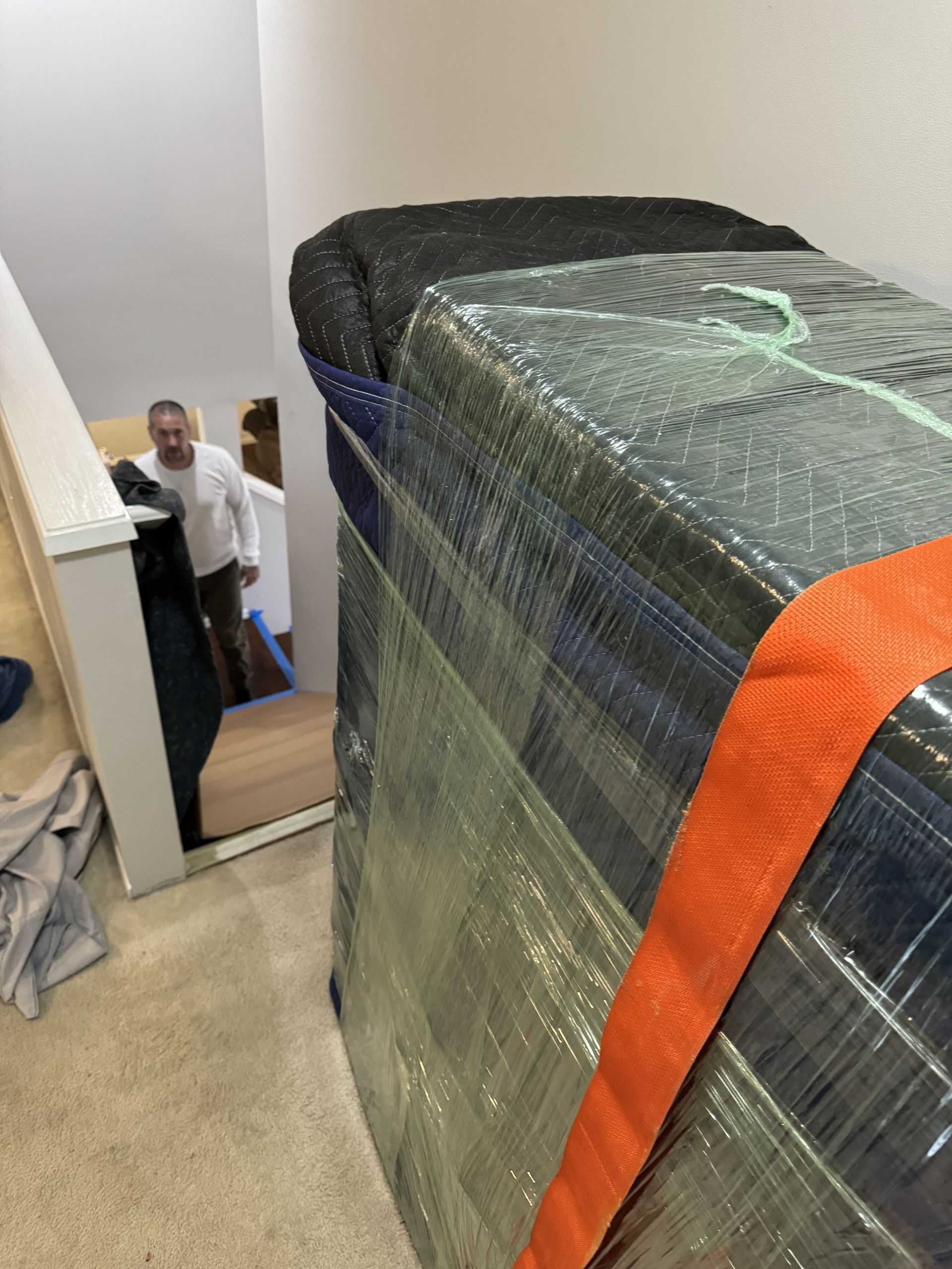 Wrapped furniture near stairs; man watches. Orange strap secures, plastic wrap covers.