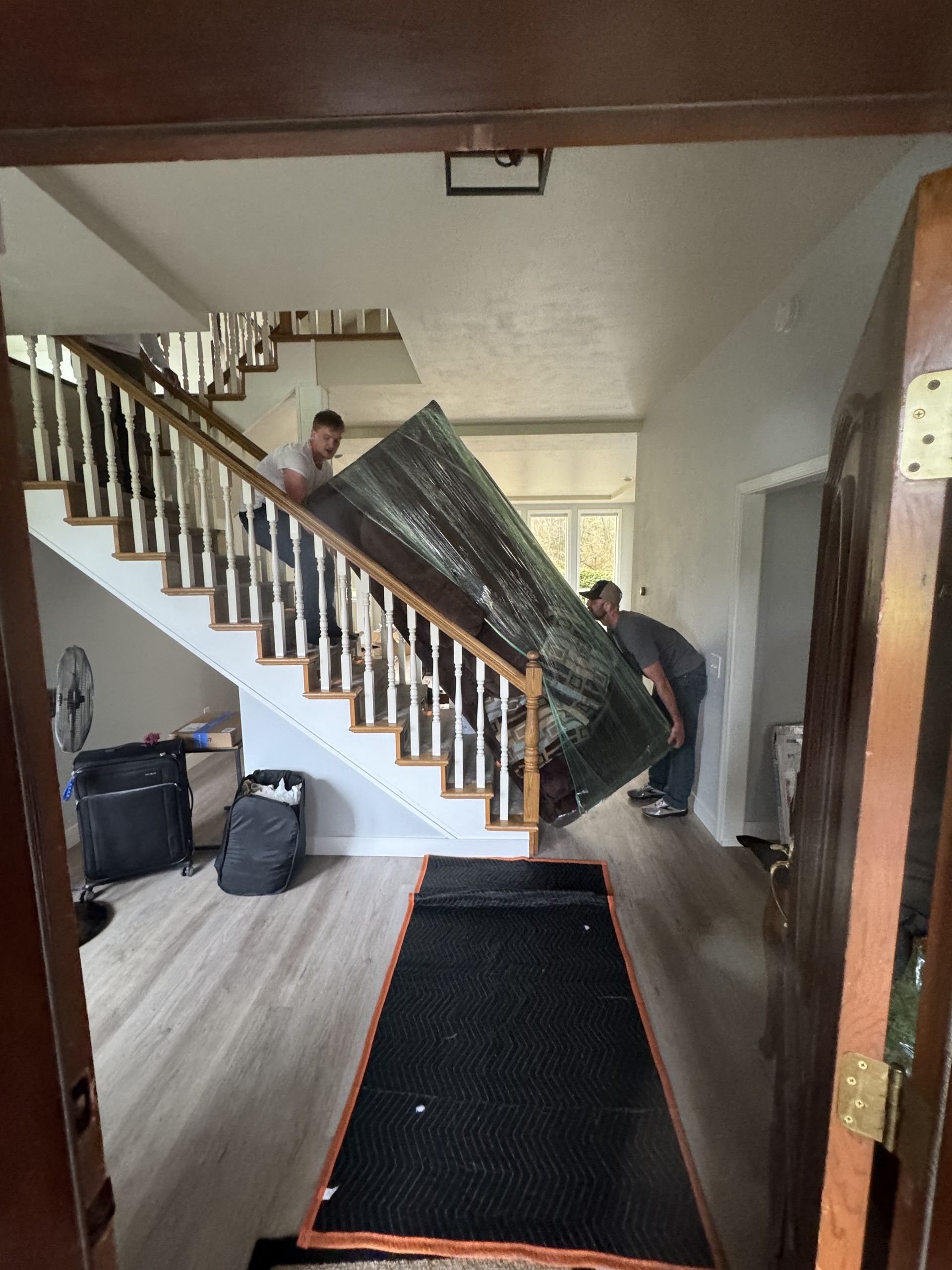 Two people carrying a large, wrapped item up a staircase inside a house.