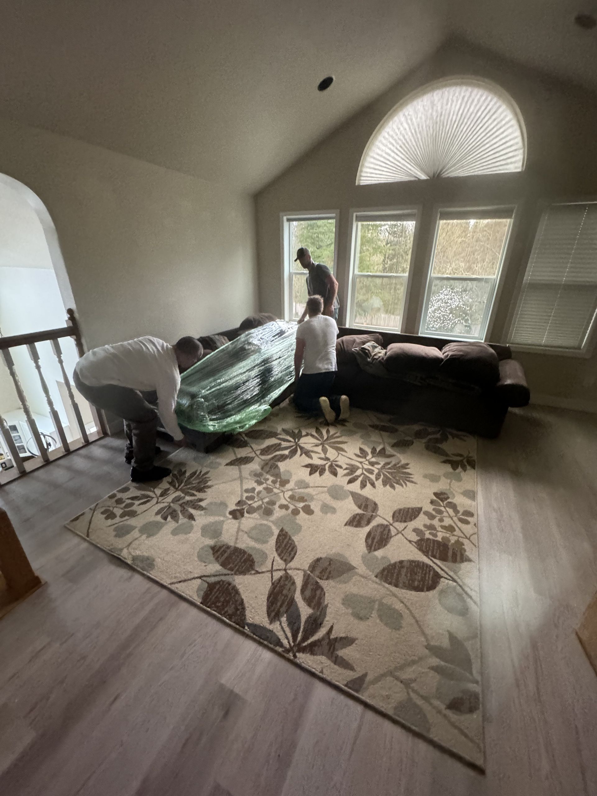 Three people moving a large, plastic-wrapped sectional sofa in a living room with a floral rug.