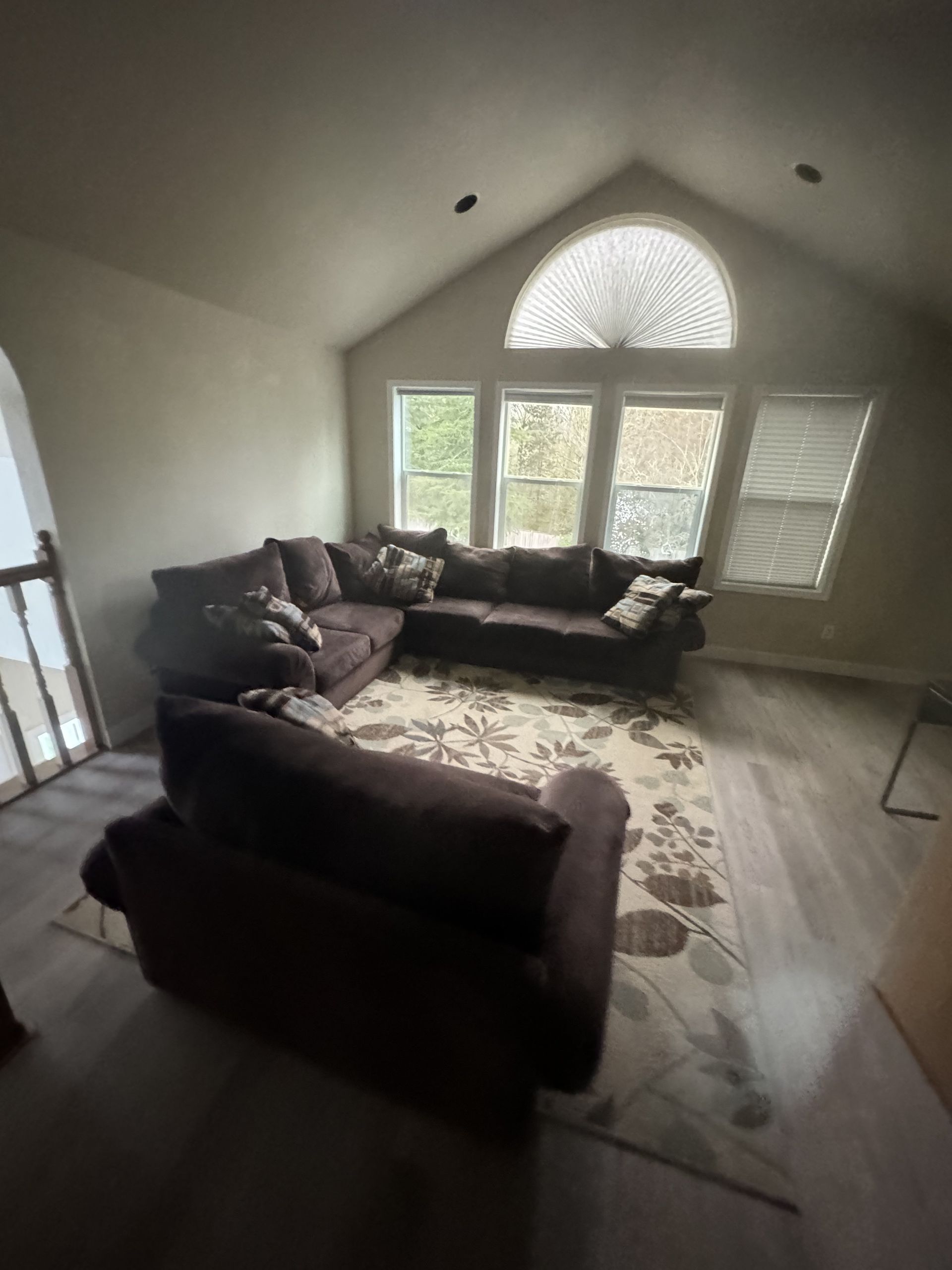 A living room with a brown sectional sofa and armchair on a floral rug, with large arched windows.
