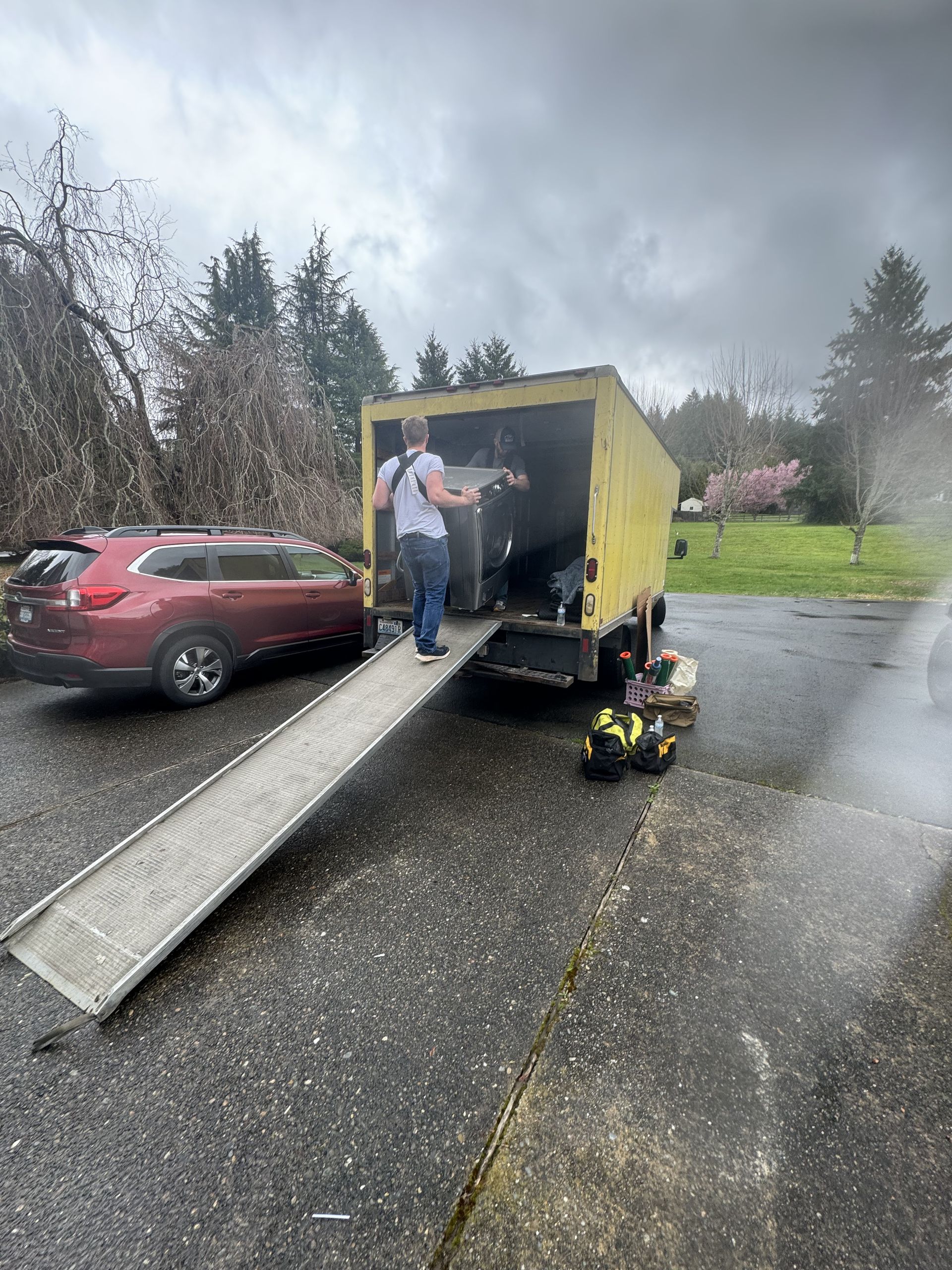 A person loading items into a yellow truck using a ramp; a red car is parked nearby. Overcast day.