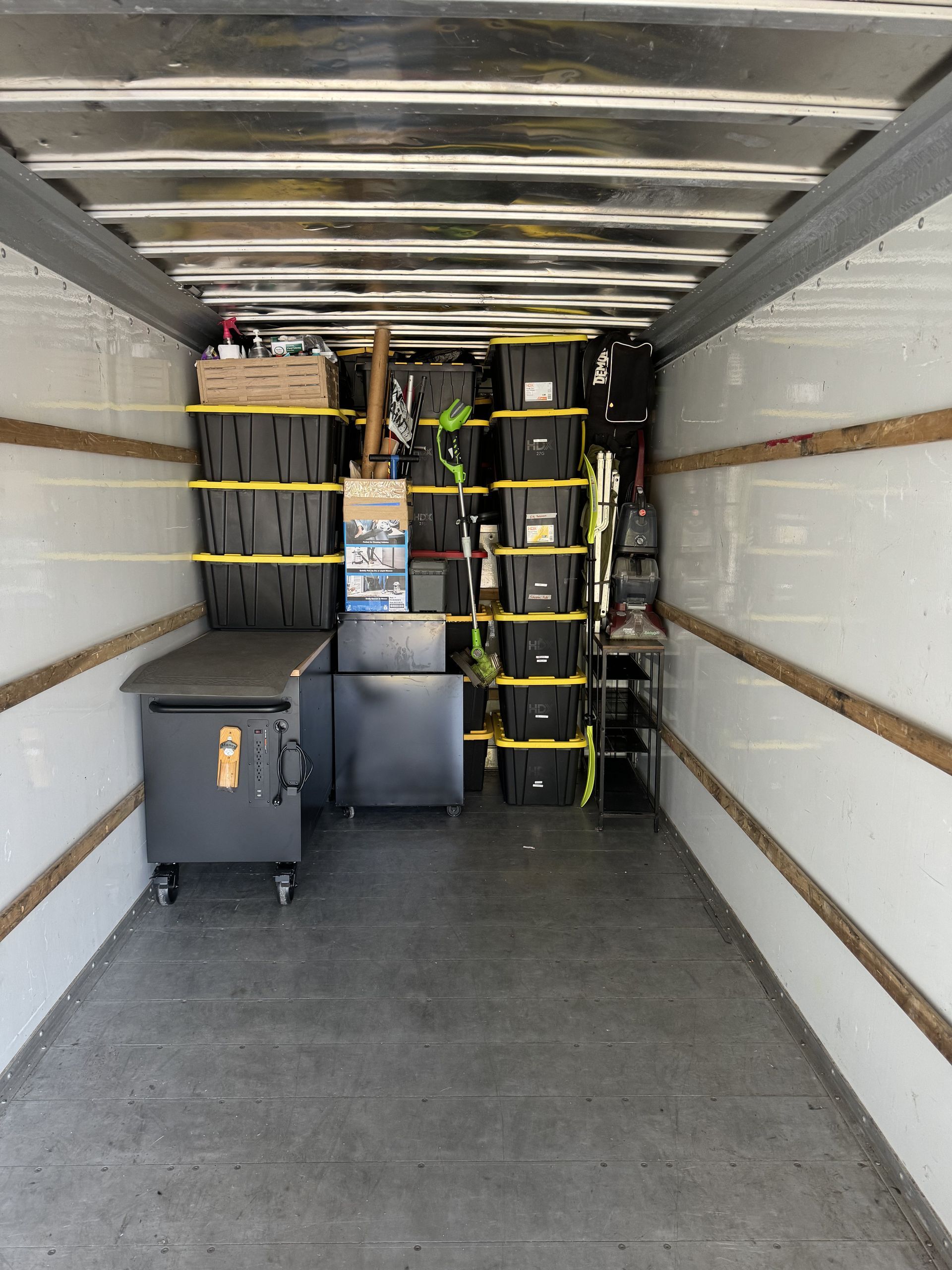 Inside of a moving truck with packed black bins, appliances, and items secured with yellow straps.