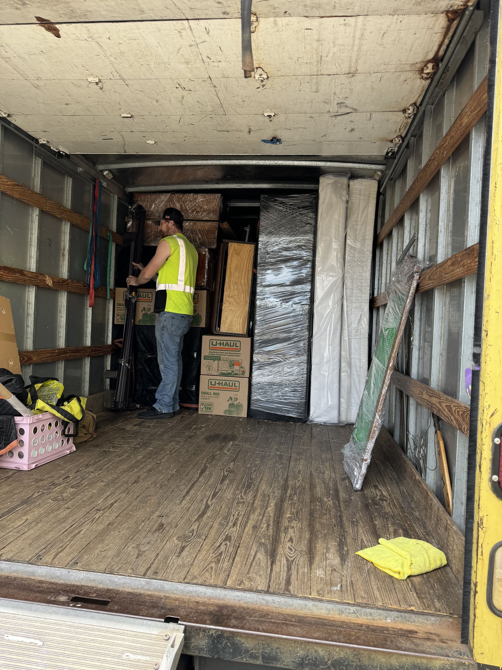 Man in safety vest loading items inside a truck, wooden walls and floor, various objects stacked.