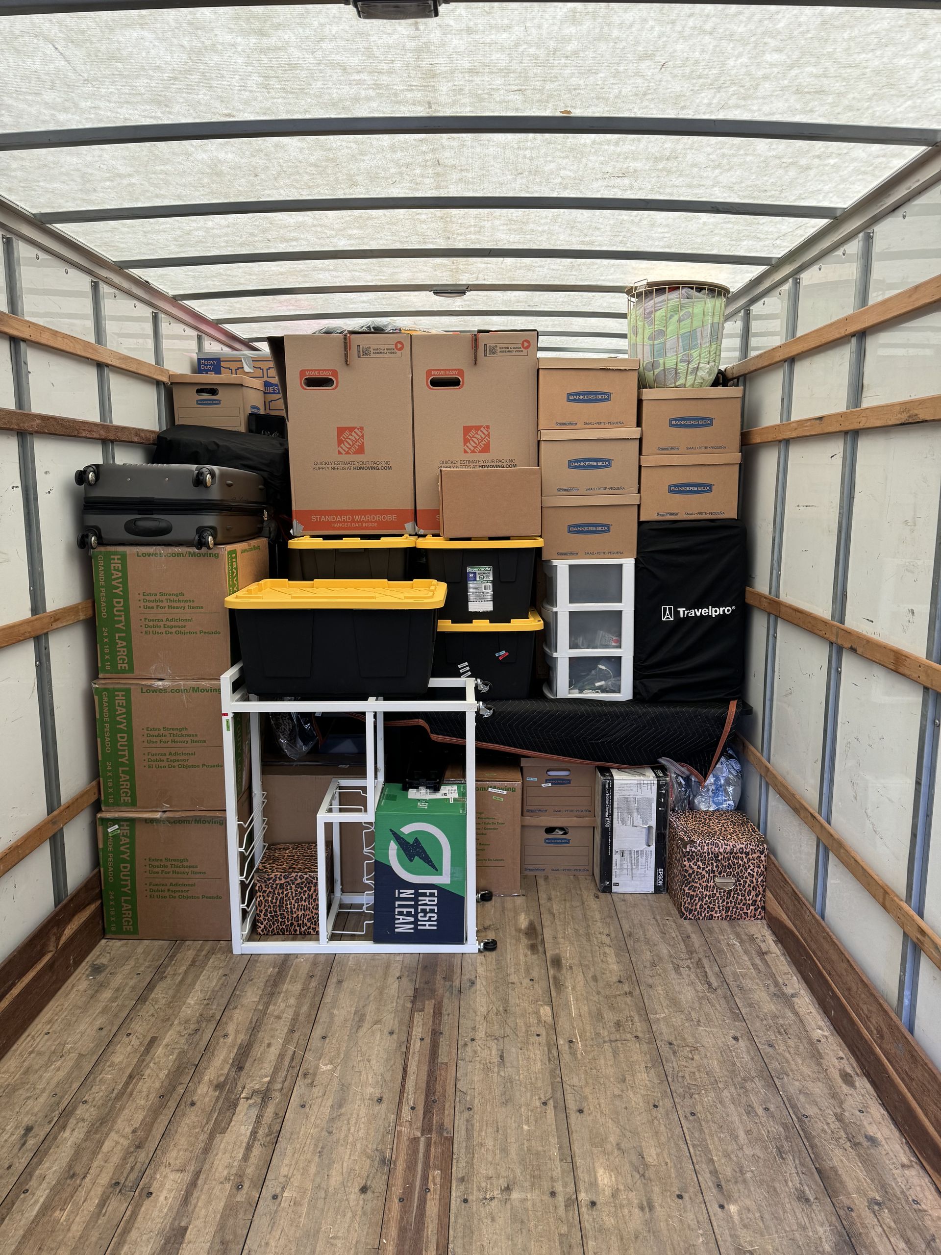 Boxes and items stacked inside a moving truck. Brown boxes, a black bin, a white appliance, and luggage fill the truck.