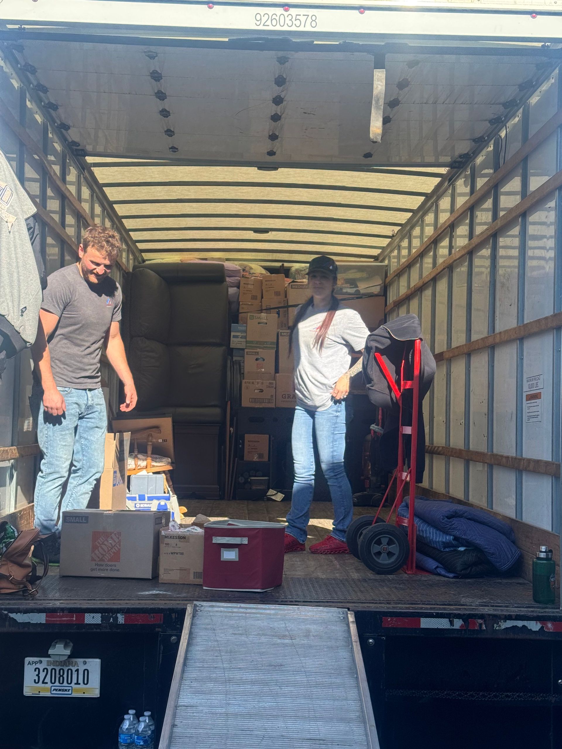 Two people inside a moving truck, loading boxes. One person stands by a hand truck. Sunny outdoor setting.