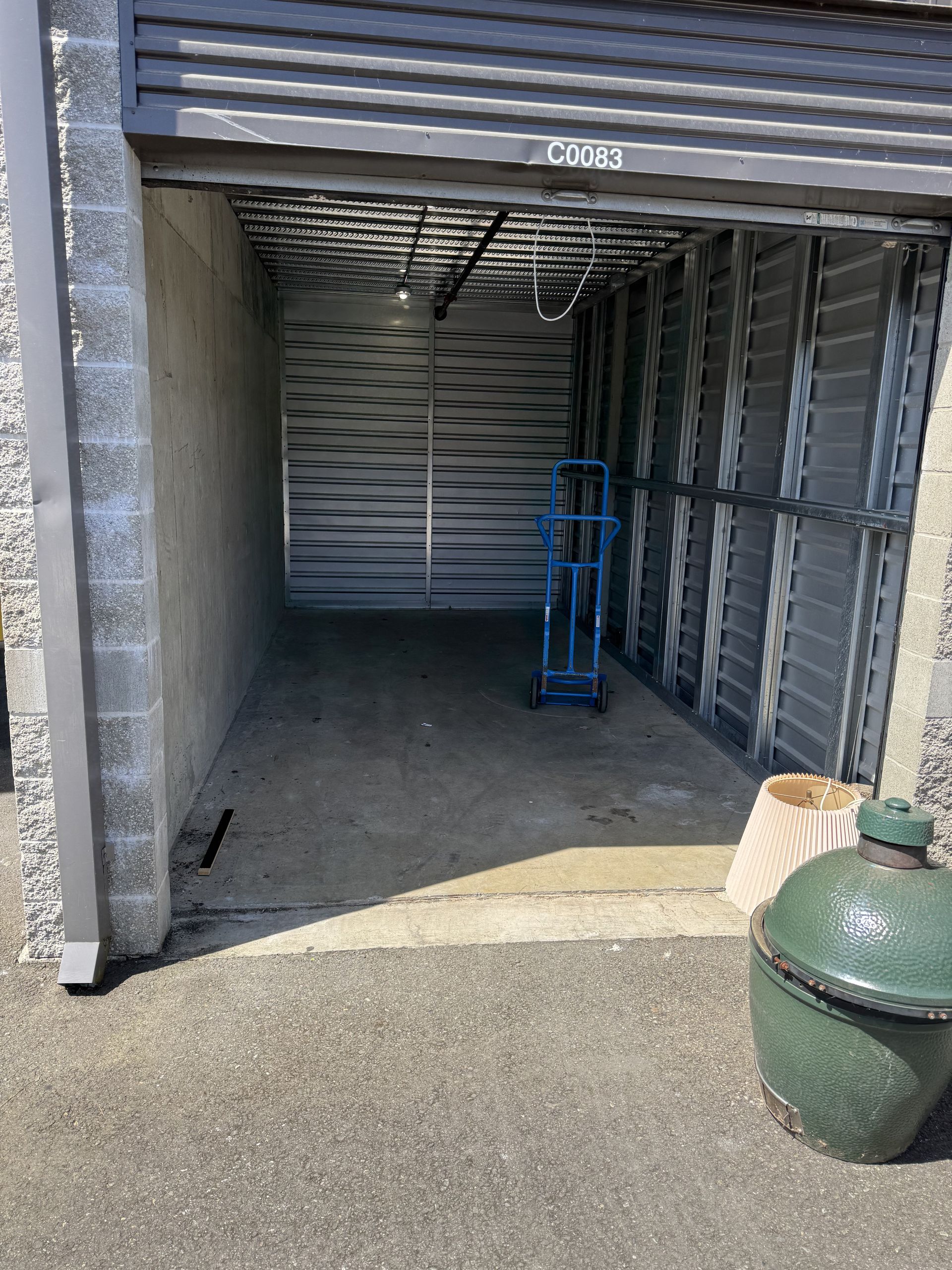 Storage unit entrance: gray roll-up door, cinder block wall, blue hand truck, green grill.