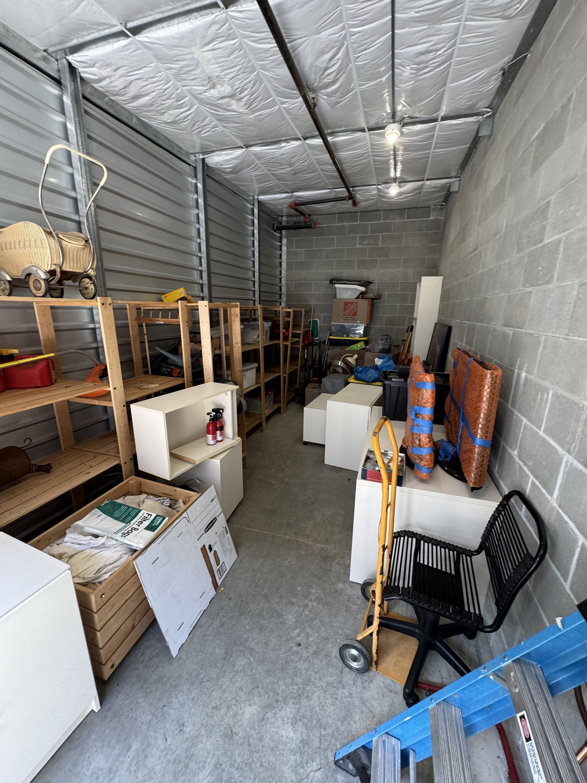 Storage unit filled with items: shelving, boxes, furniture, and a cart. Gray walls and ceiling.