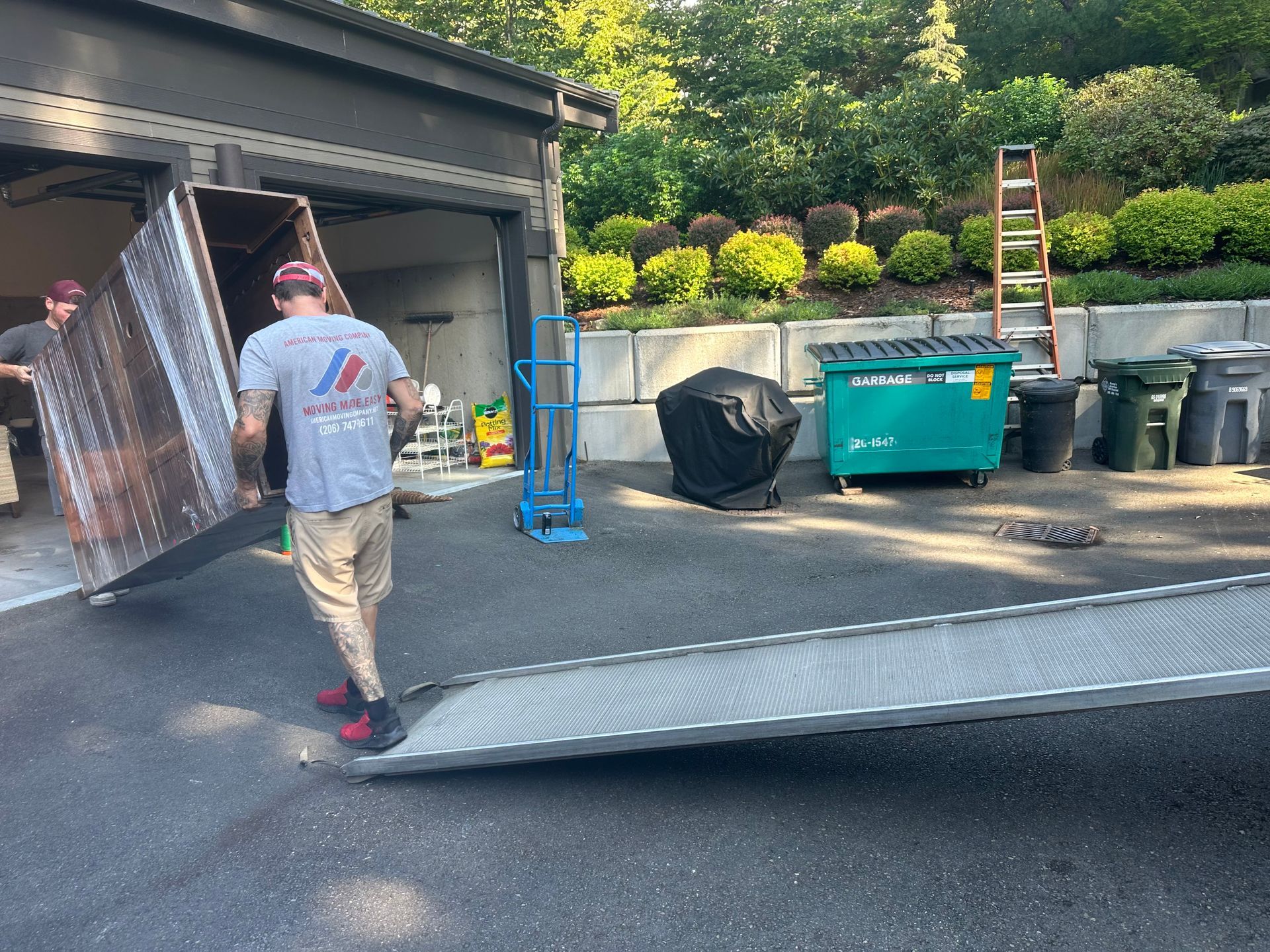 Two movers carrying a large panel out of a garage onto a ramp. Green dumpster and shrubbery in the background.