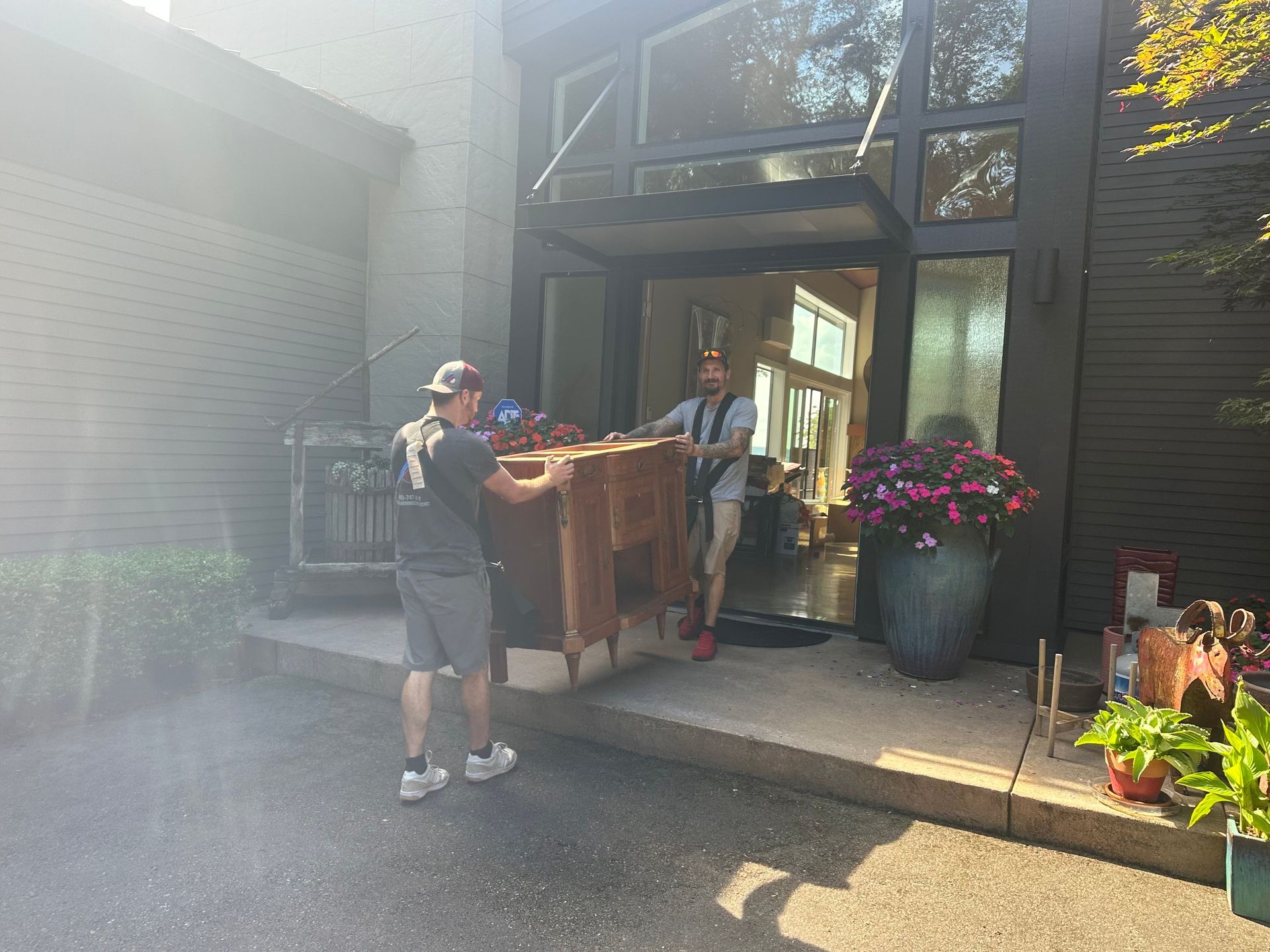 Two movers carrying a large wooden cabinet out of a modern home with glass doors.