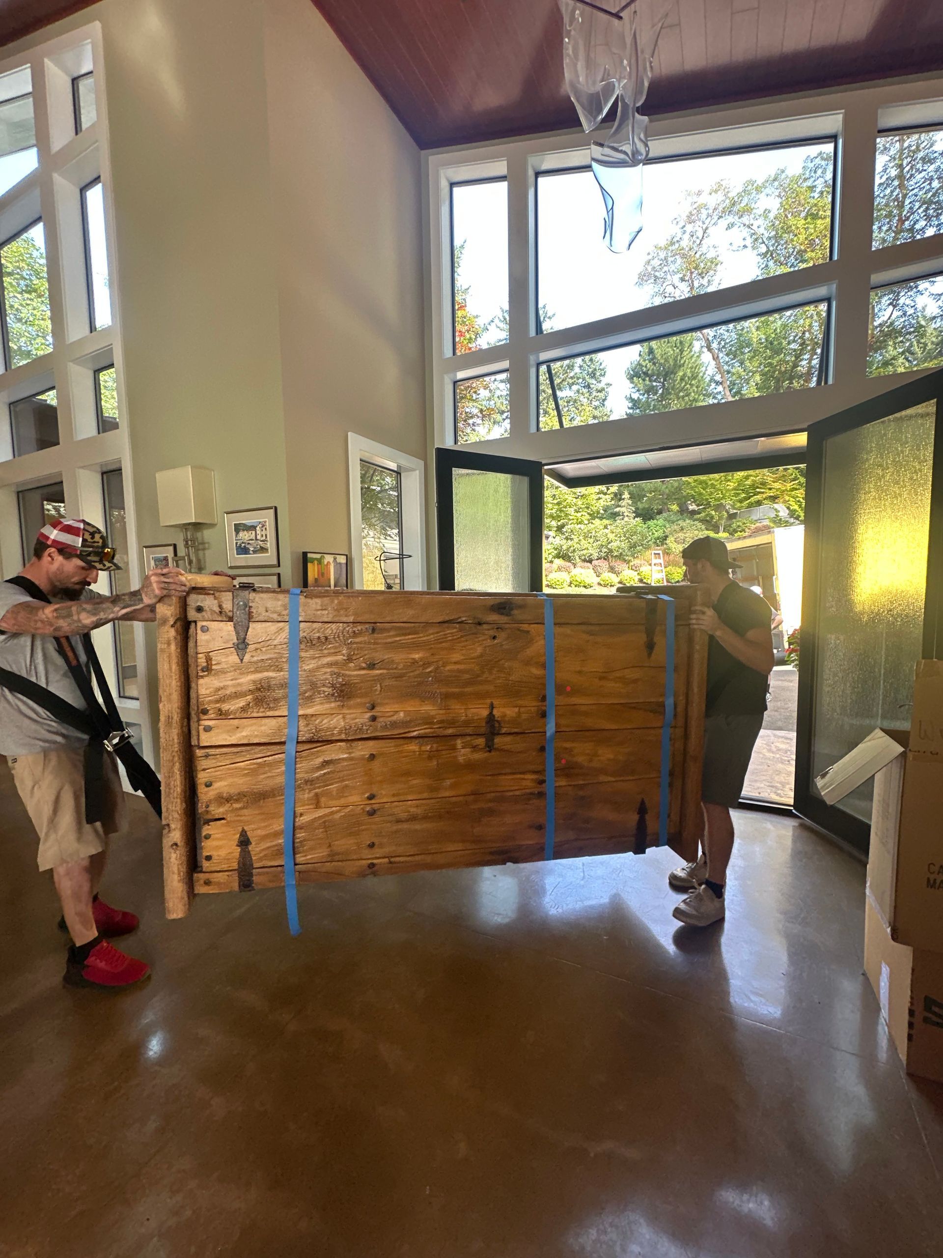 Two people carrying a large, wooden crate through an open doorway. Interior with large windows and trees visible outside.