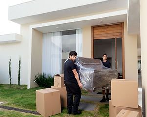Two movers carrying a wrapped sofa out of a house with boxes on the lawn.