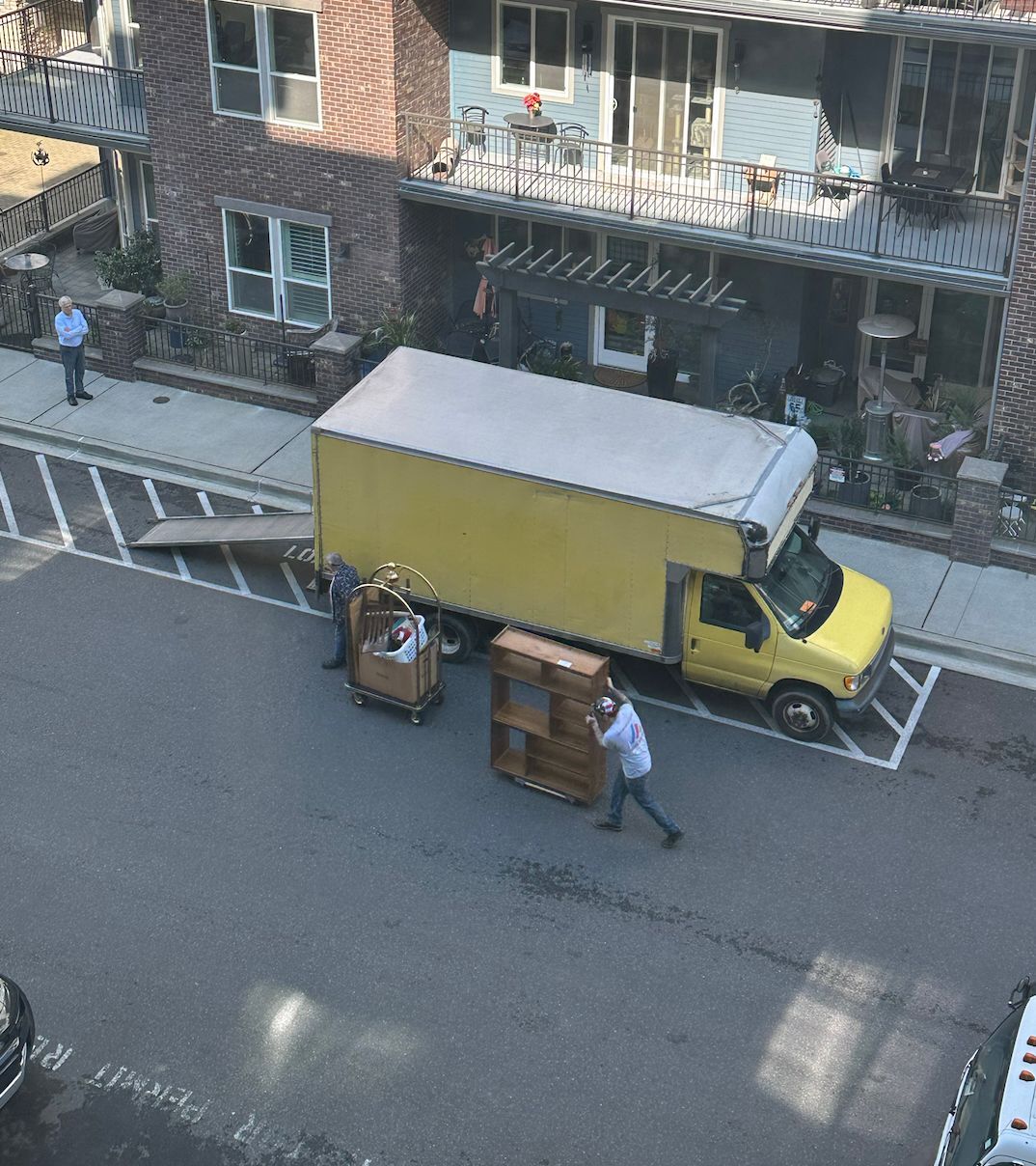 Movers loading furniture onto a yellow moving truck parked on a street near a brick building.