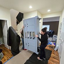 People moving a tall, gray cabinet through a hallway. They are wearing casual clothing.