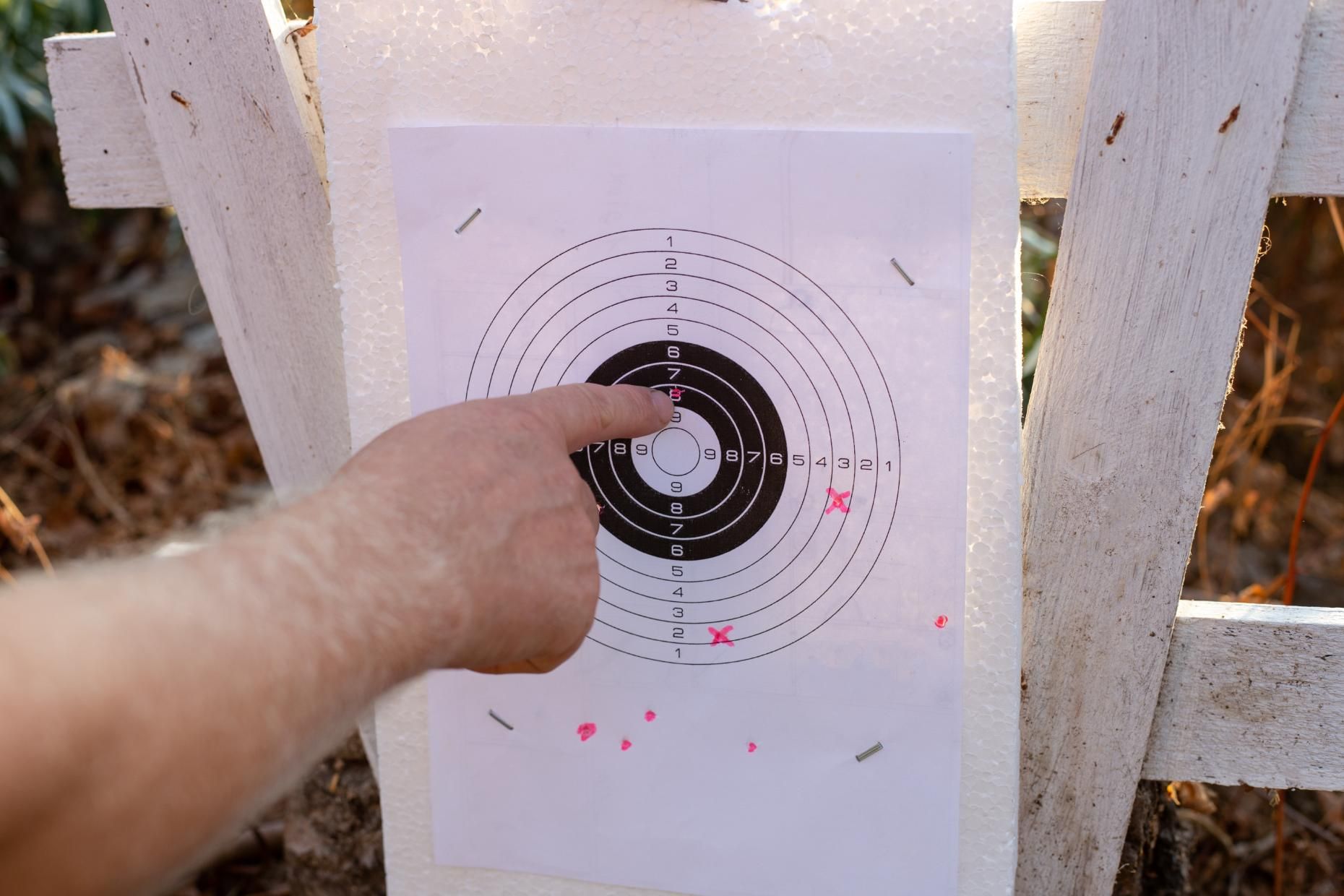 A person's finger pointing at a target with bullet holes. The target is centered on a white board.