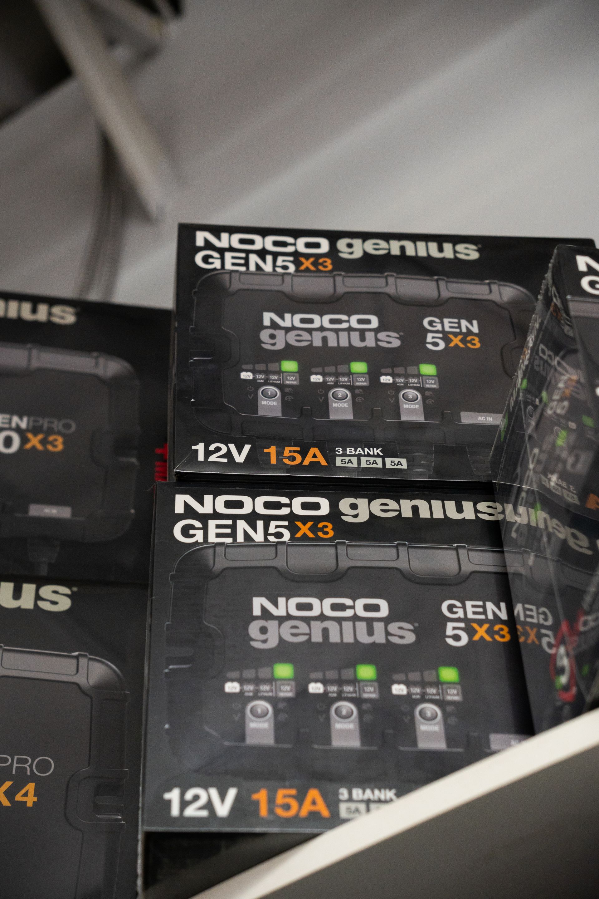 Des boîtes de chargeurs de batterie NOCO Genius GEN5X3 sont rangées sur une étagère. Les boîtes sont noires avec des inscriptions blanches et des indicateurs LED verts.