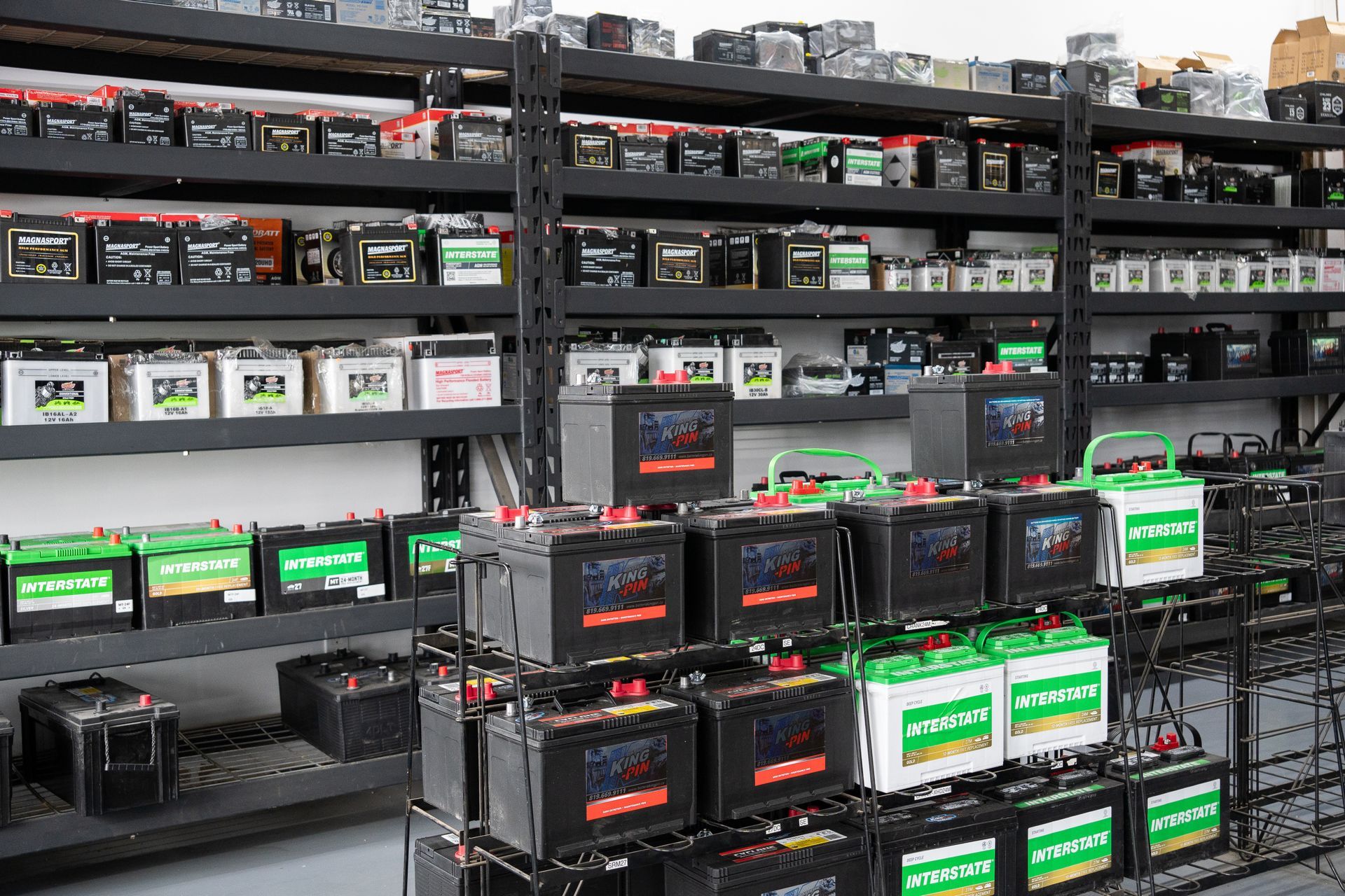 Des rangées de batteries de voiture dans un magasin, des étagères en métal noir, de marques et de couleurs variées.