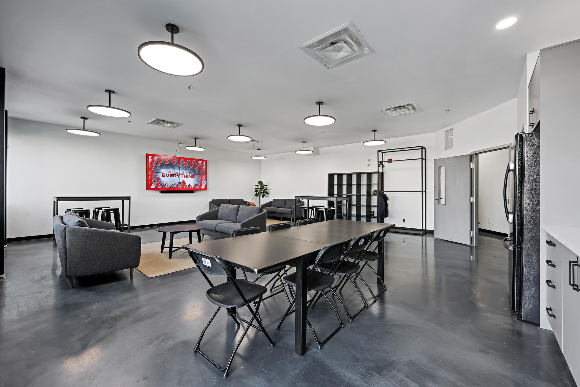 Modern lounge area featuring a large table with black chairs, gray sofas, a wall-mounted TV, and polished concrete floors.
