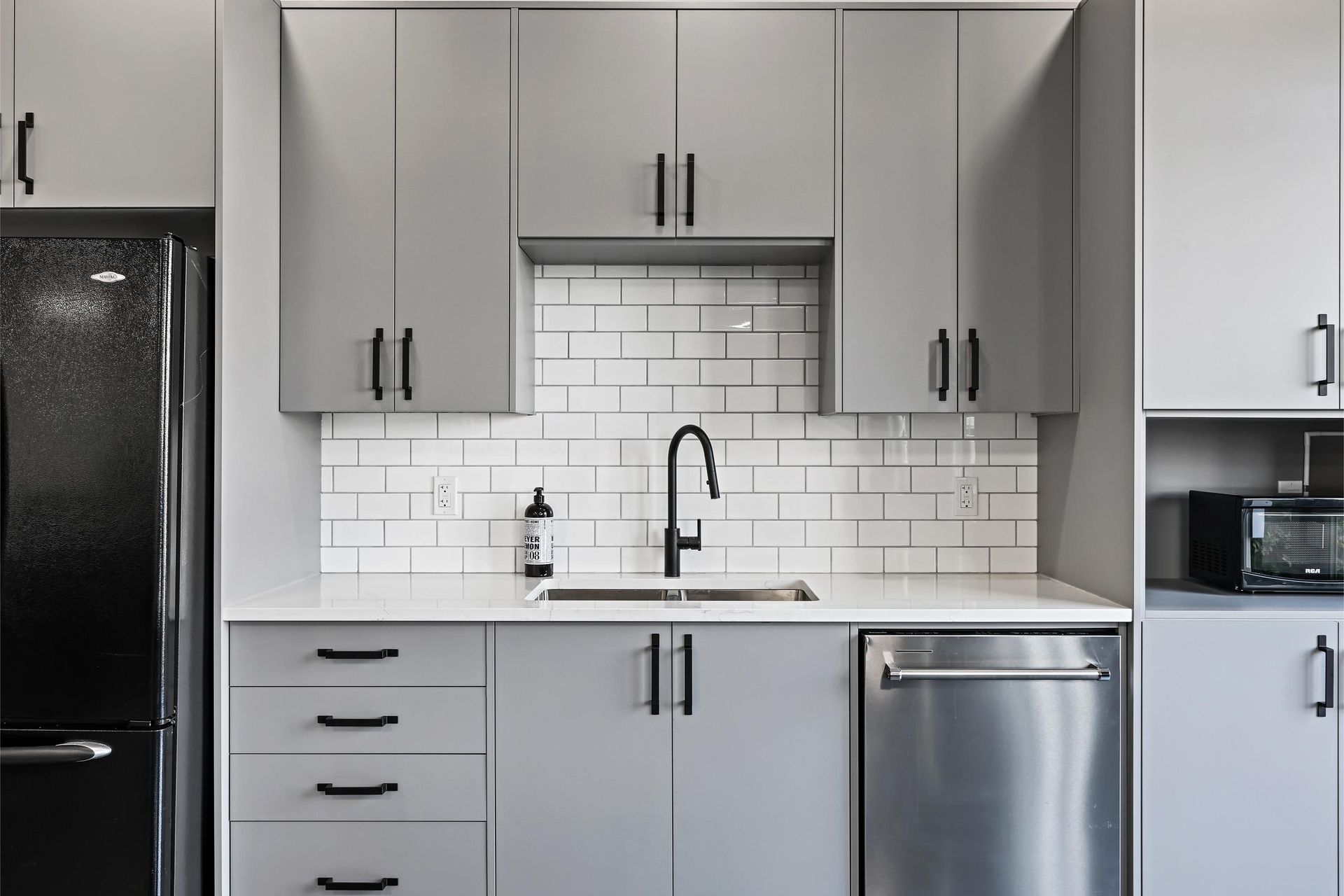 A modern kitchen featuring gray cabinets, a white subway tile backsplash, a stainless steel dishwasher, and a black sink.