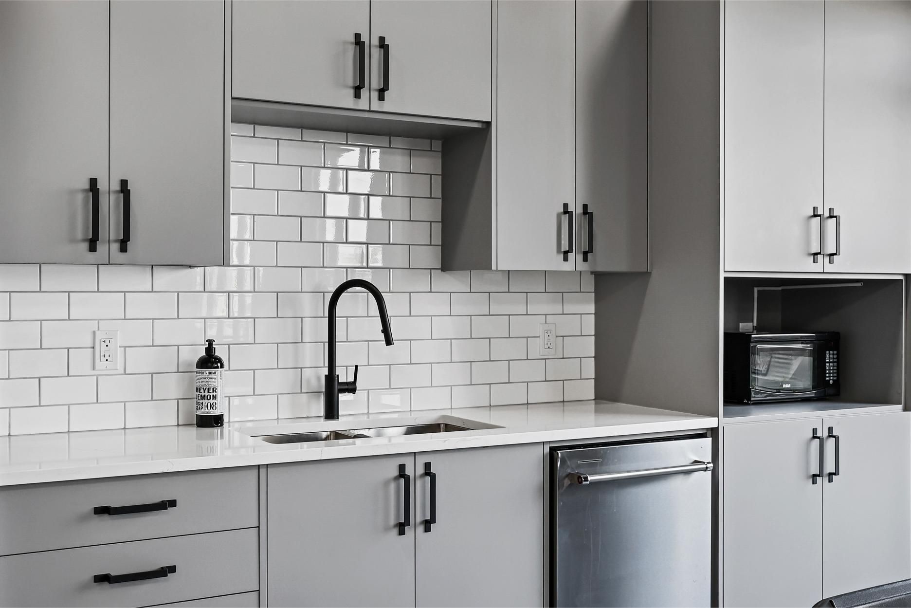 A modern kitchen featuring matte gray cabinets, a white subway tile backsplash, a black faucet, and stainless appliances.