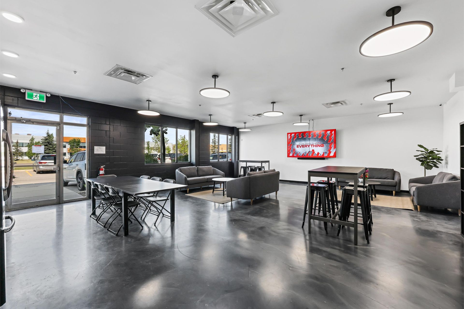 A modern, spacious office lounge featuring gray floors, contemporary furniture, pendant lighting, and large windows.