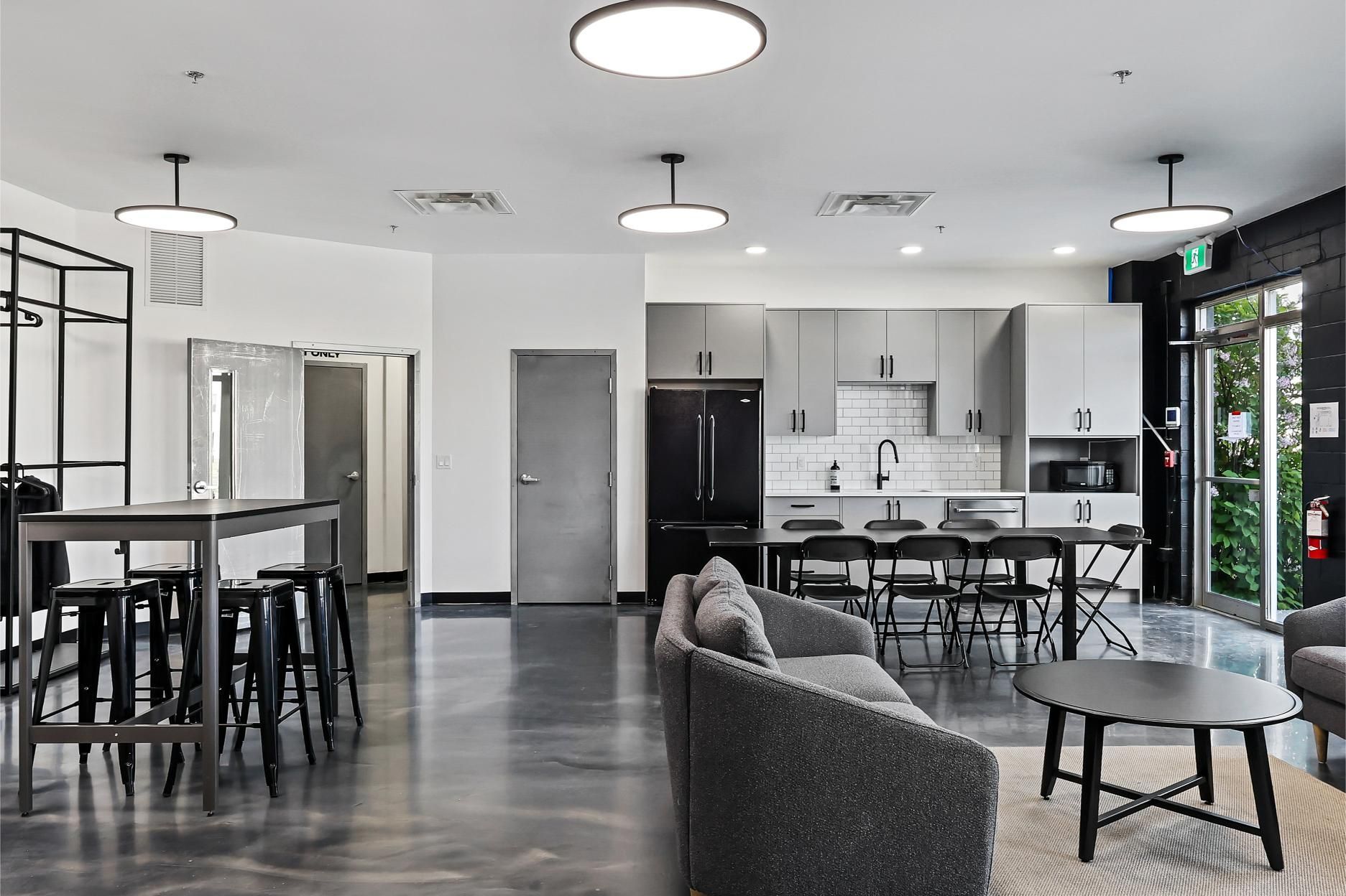 Modern breakroom with a small kitchen, seating area, and tall dining table on a glossy concrete floor.