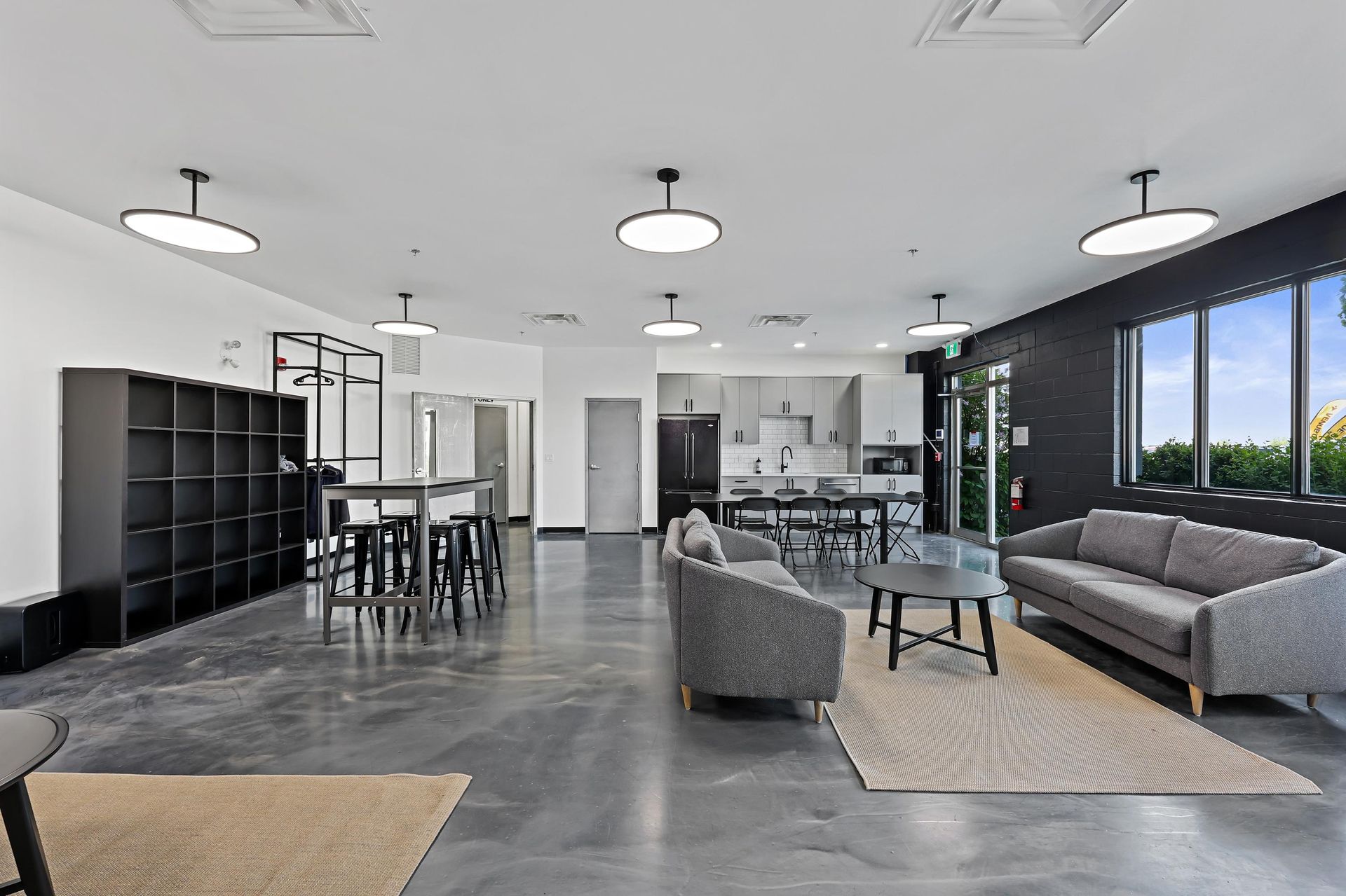 A modern lounge area with gray sofas, black shelving, a high-top table, and a small kitchenette on concrete floors.