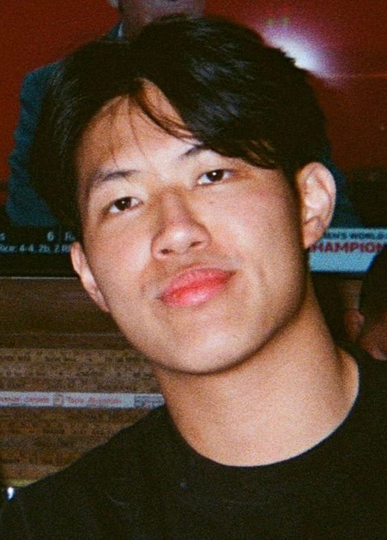 Person with dark hair, looking at the camera, slight smile. Indoors, blurred background.