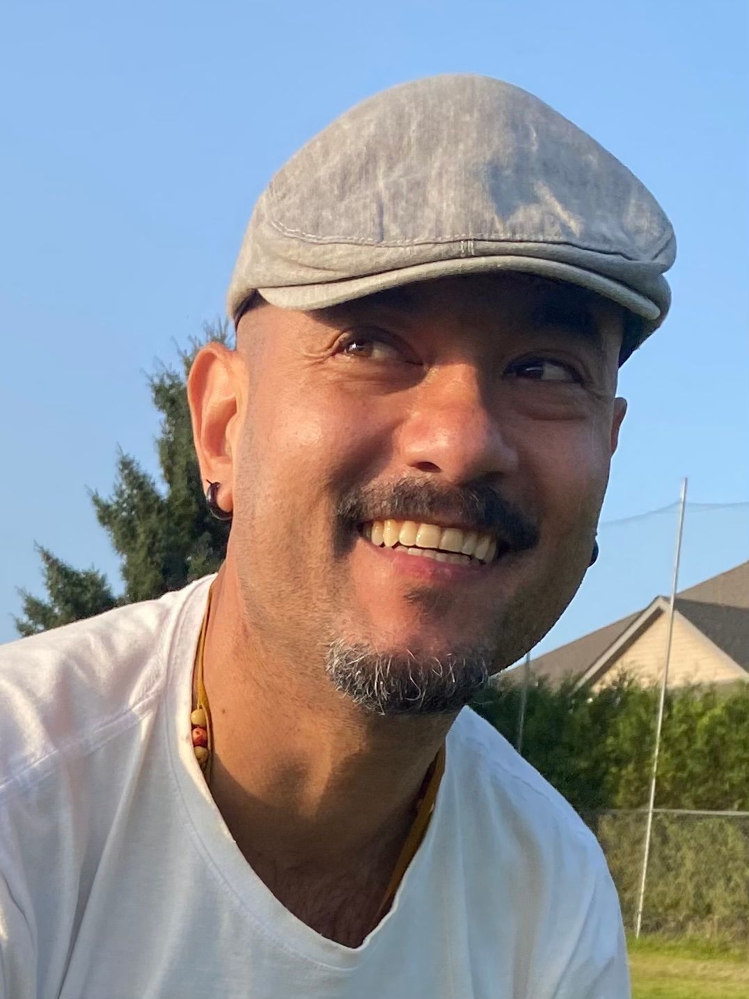 Person with a gray cap smiling, outdoors. White t-shirt, small beard, earring. Blue sky, green grass in background.