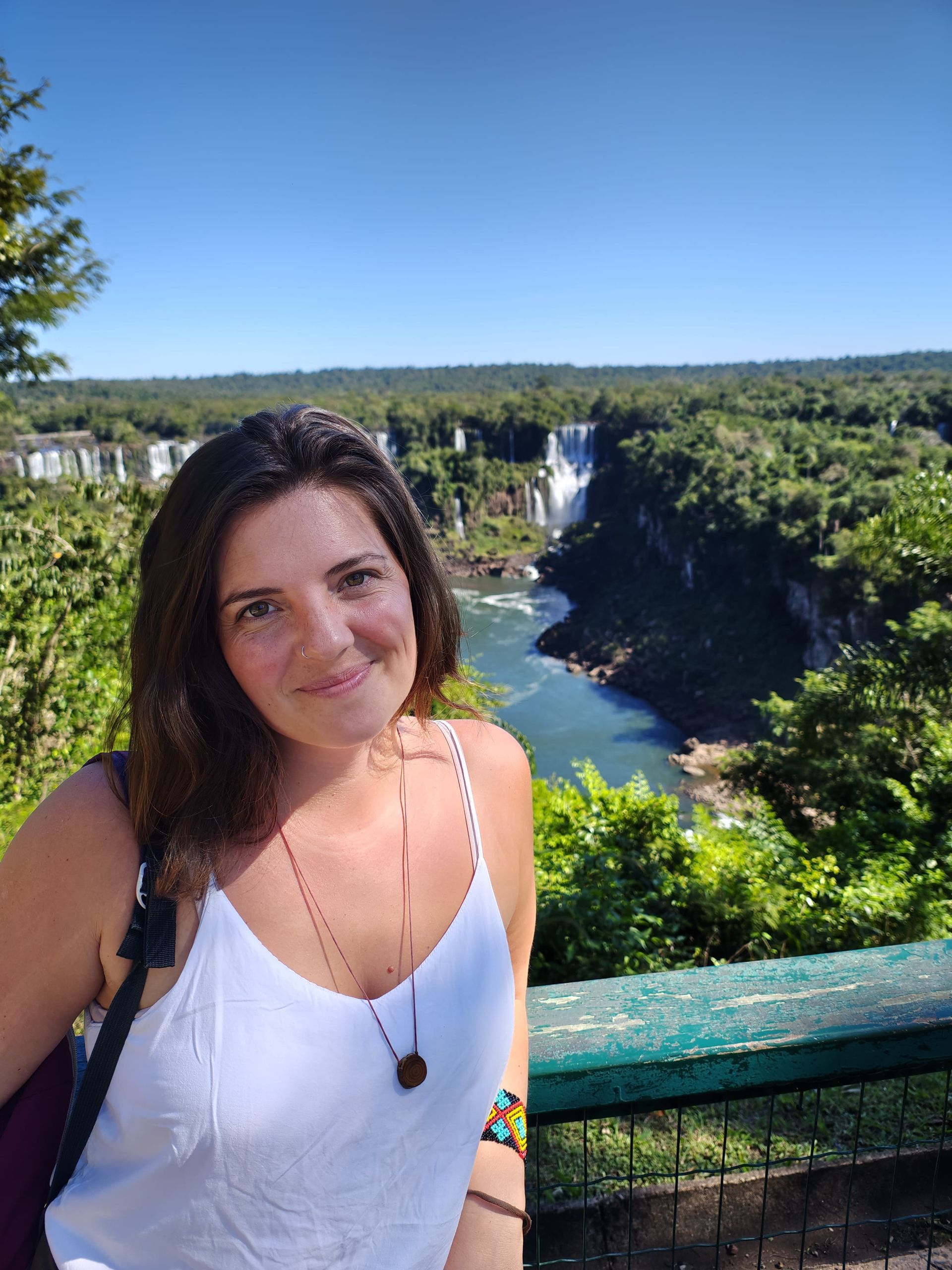 Person in white tank top smiles at camera with waterfalls and lush green landscape in background.