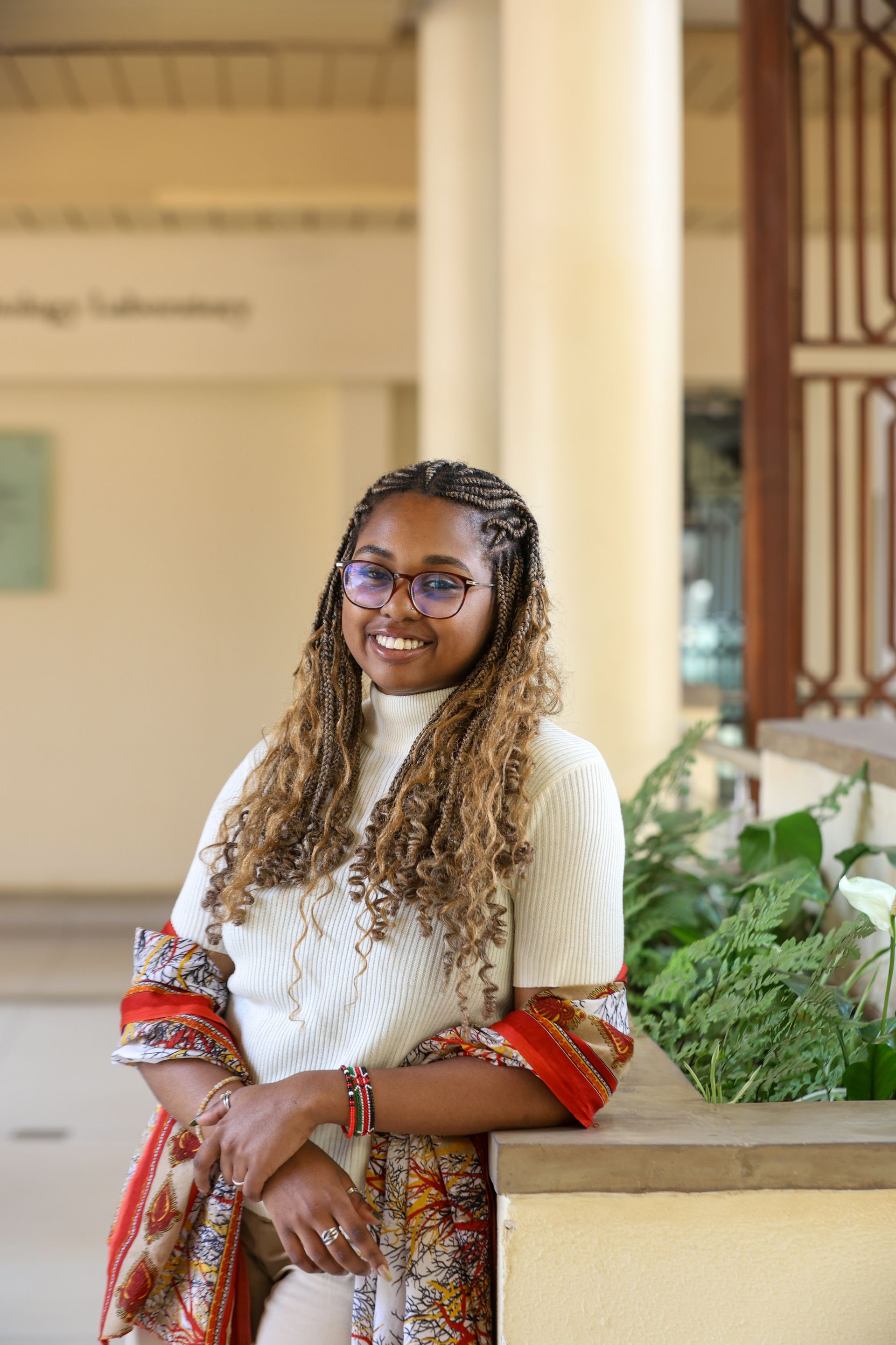 Person with glasses, braids, wearing a patterned scarf and sweater, smiling and leaning on a stone ledge near a column.